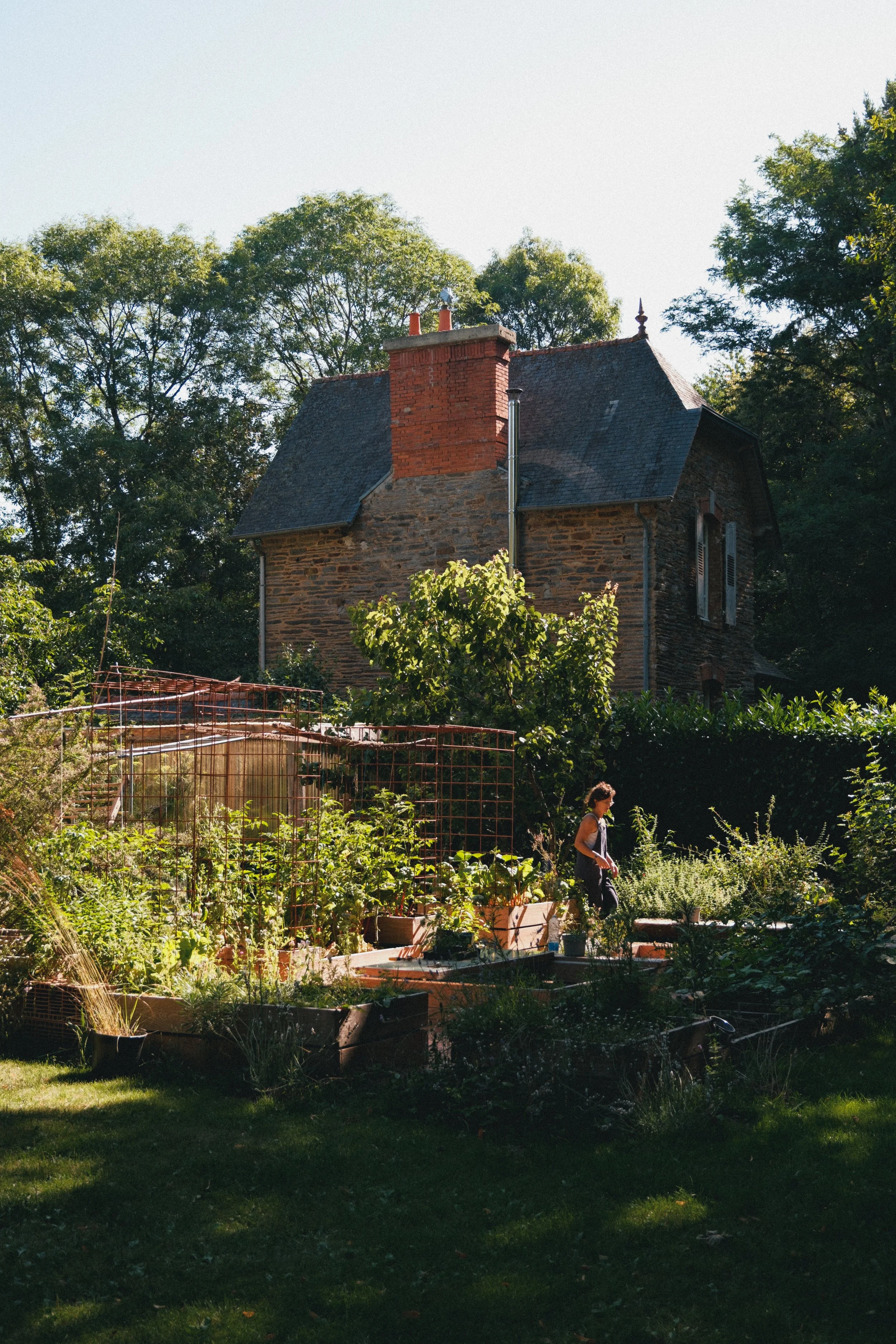 Une maison en pierre avec un toit en ardoise, entourée de végétation abondante et un jardin potager. Une femme marche dans le jardin.