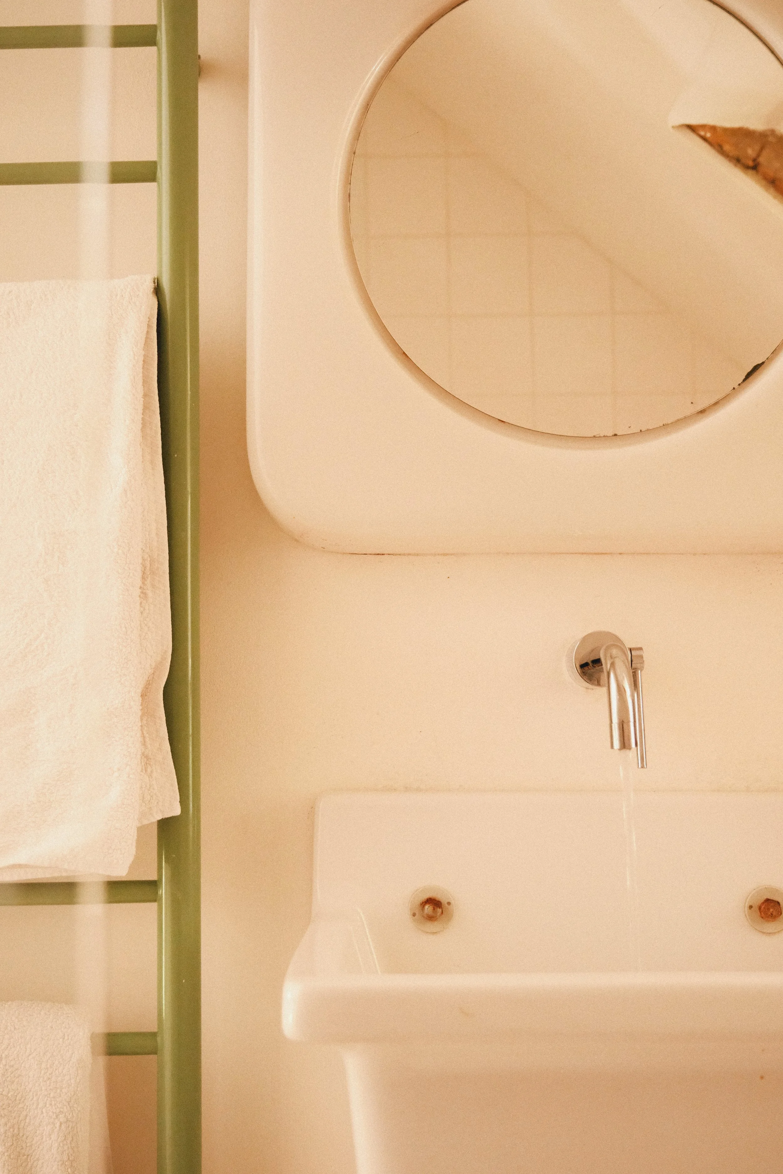 Une salle de bain avec un lavabo blanc, un miroir rond, un robinet en métal, un porte-serviettes vert, une serviette blanche, et des taches de rouille dans l'évier.