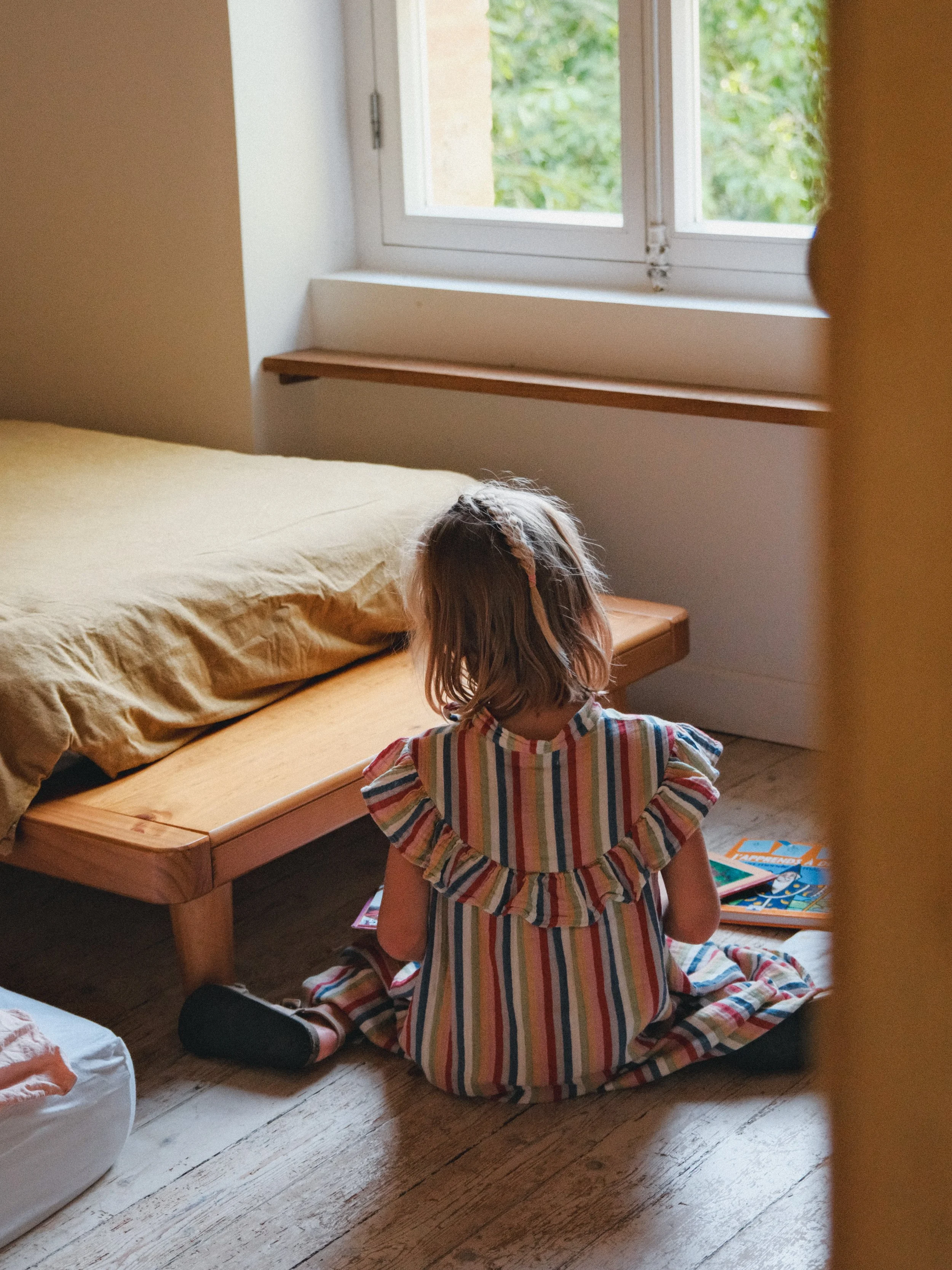 Une jeune fille avec des cheveux longs, vêtue d'une robe rayée de couleurs vives, assise par terre près d'un lit en bois, regardant un livre ou un jeu de société. La scène se déroule dans une pièce lumineuse avec une fenêtre laissant entrer la lumièr