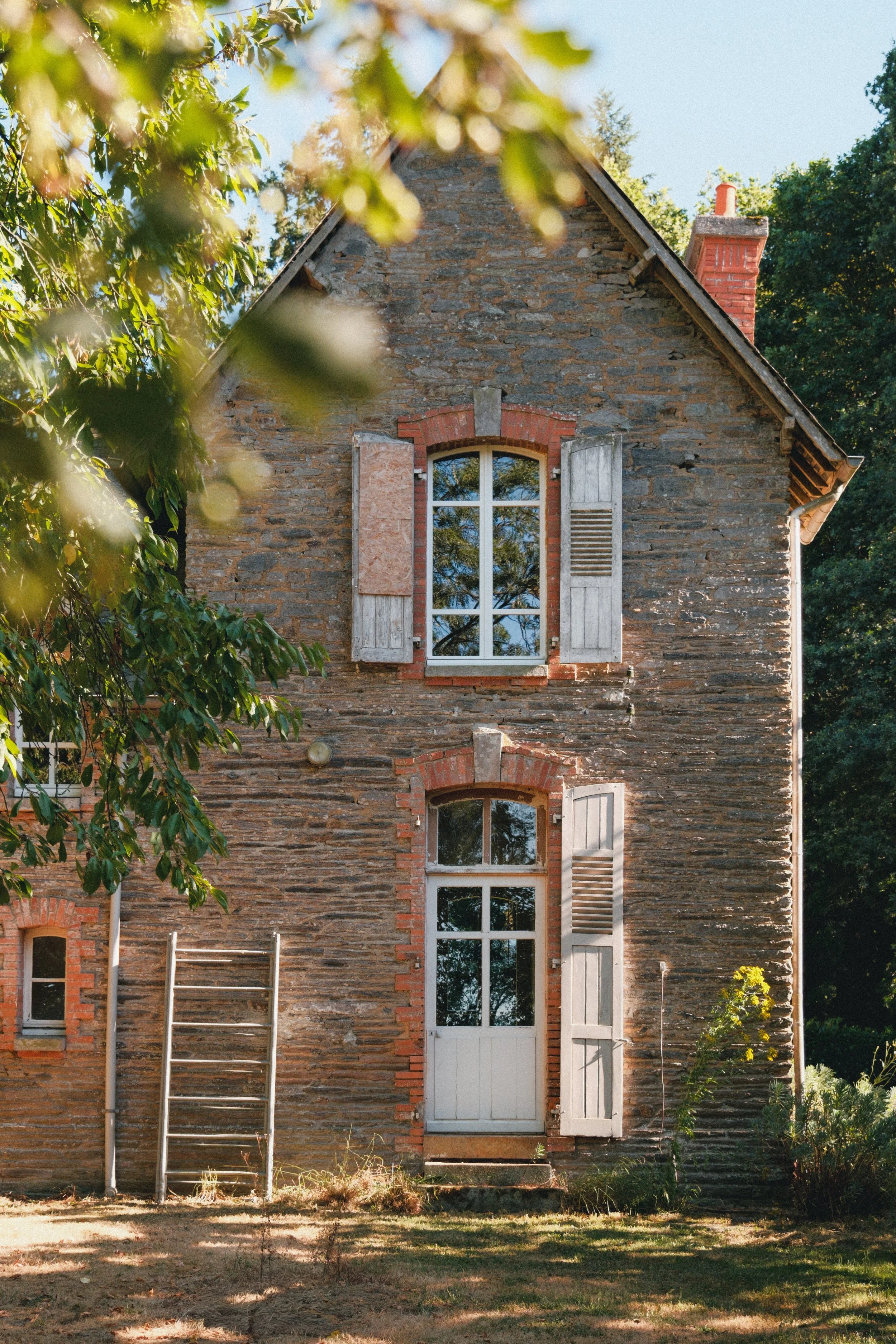 Maison en pierre avec fenêtres à volets en bois, entourée de végétation verte, avec un ciel bleu en arrière-plan.