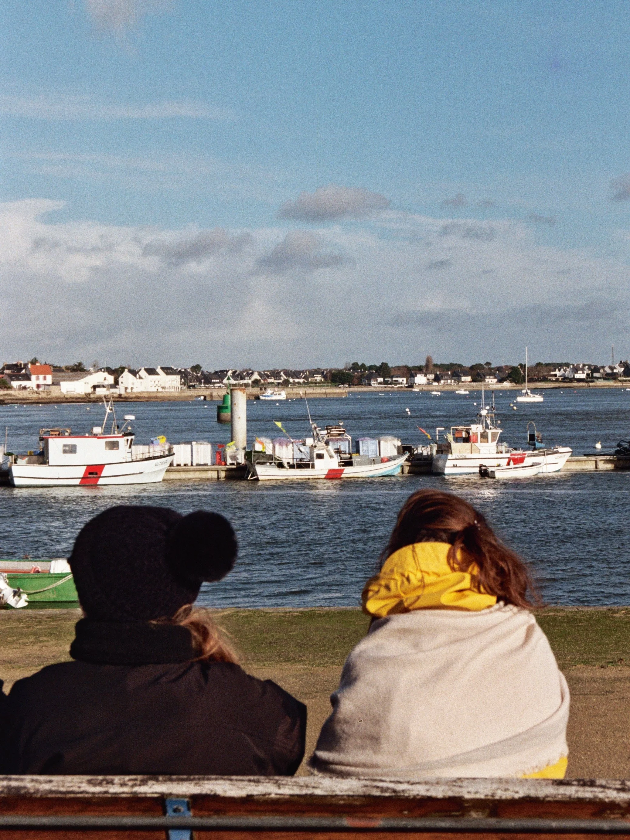 Vue de trois personnes assises à un bord de mer, regardant des bateaux sur l'eau avec des maisons en arrière-plan et un ciel partiellement nuageux.