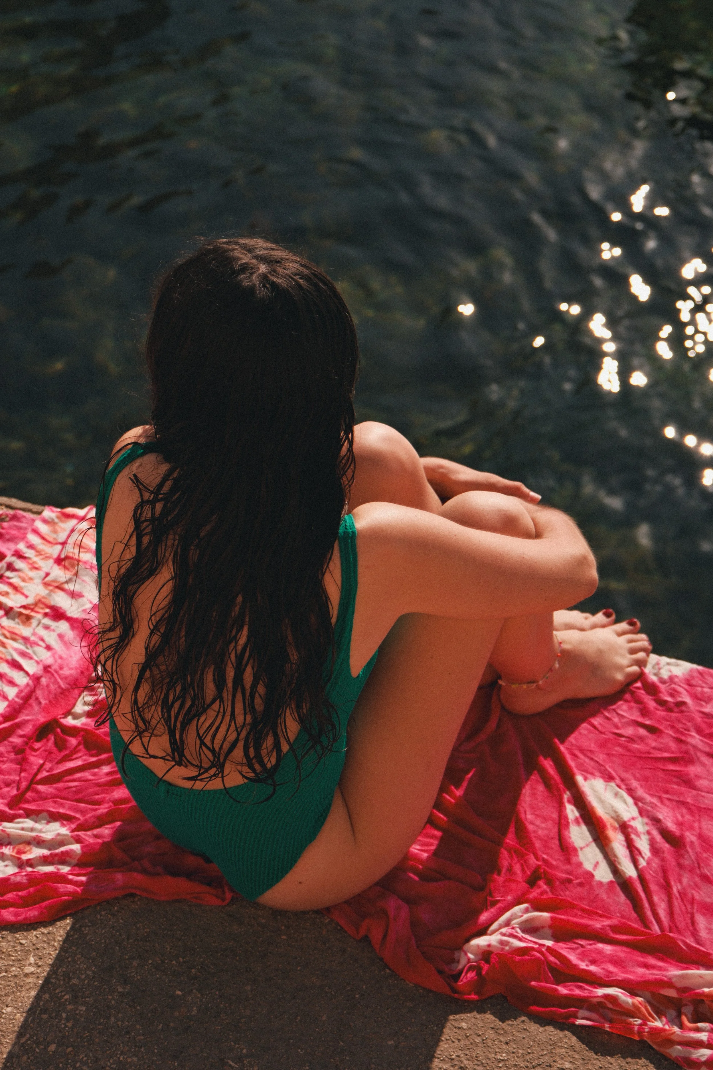 Une femme assise sur un tapis rouge près de l'eau, portant un maillot vert foncé, avec des cheveux longs et mouillés, regardant vers le lac ou la rivière.