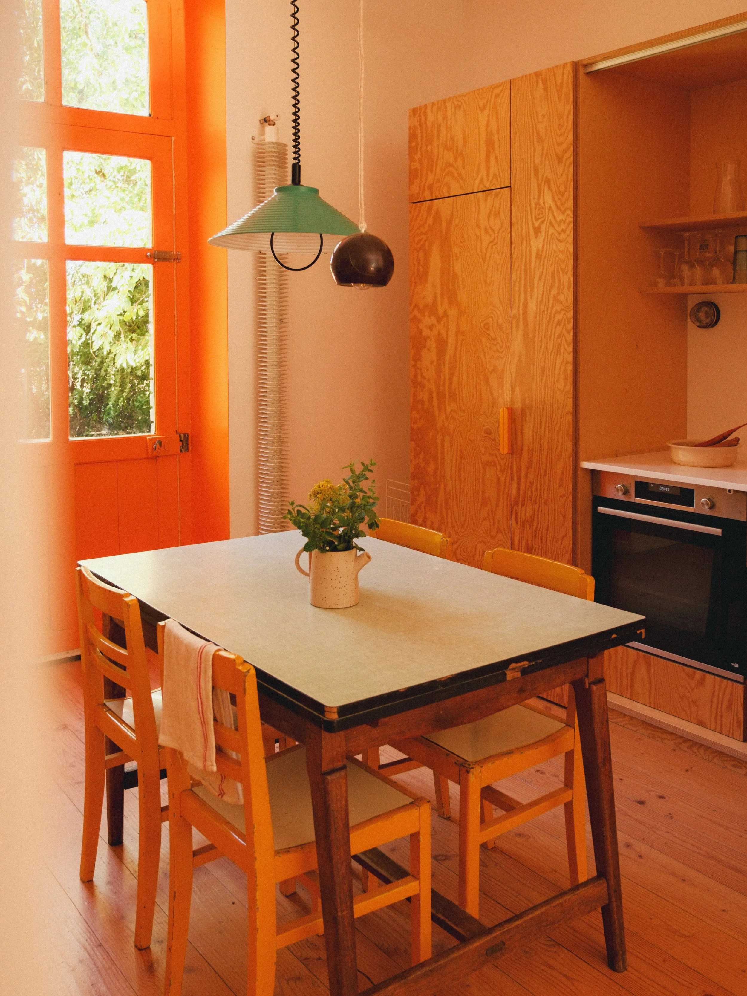 Une cuisine en bois avec une table en bois, des chaises en bois, une fenêtre avec cadre orange, une lampe suspendue verte, des étagères avec des verres et une assiette, un four encastré.