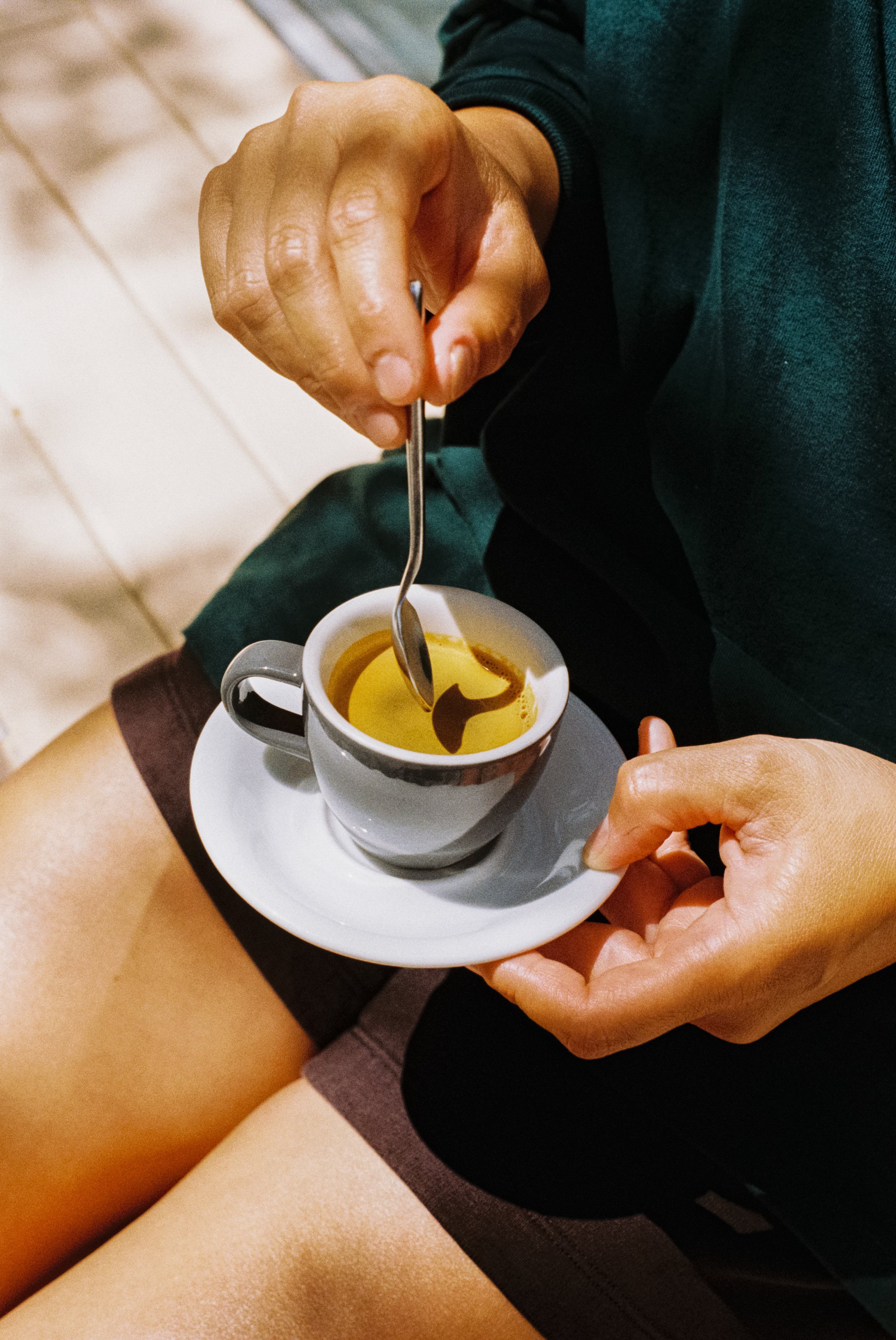 Une personne tient une cuillère à café dans sa main, en train de verser du caramel dans une tasse de café. La tasse est posée sur une assiette blanche, et la personne a l'autre main sur la tasse. La personne porte un vêtement noir et est assise sur u