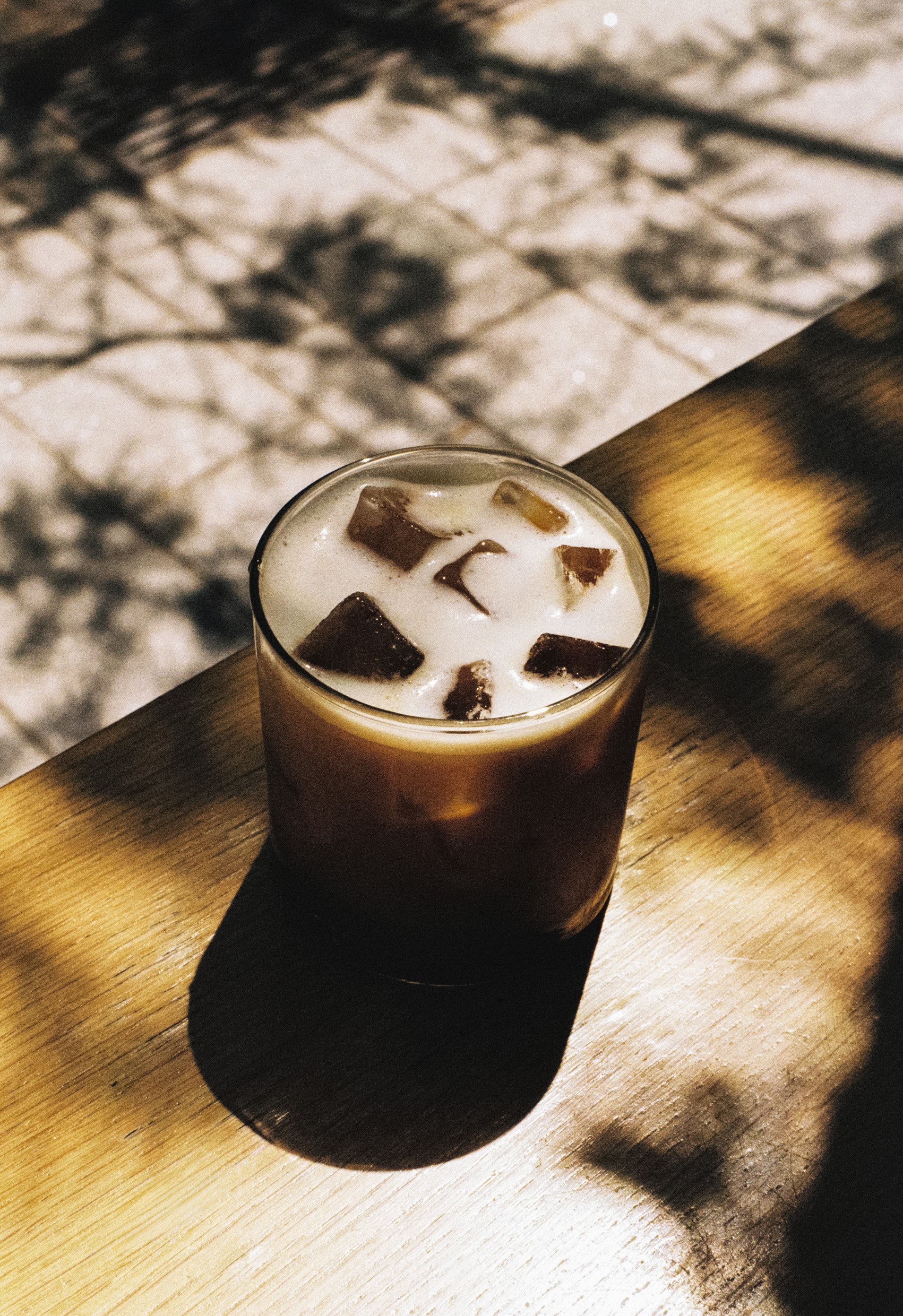 Un verre de café glacé avec des morceaux de glace sur une surface en bois, avec un motif d'ombres de grille sur le fond.