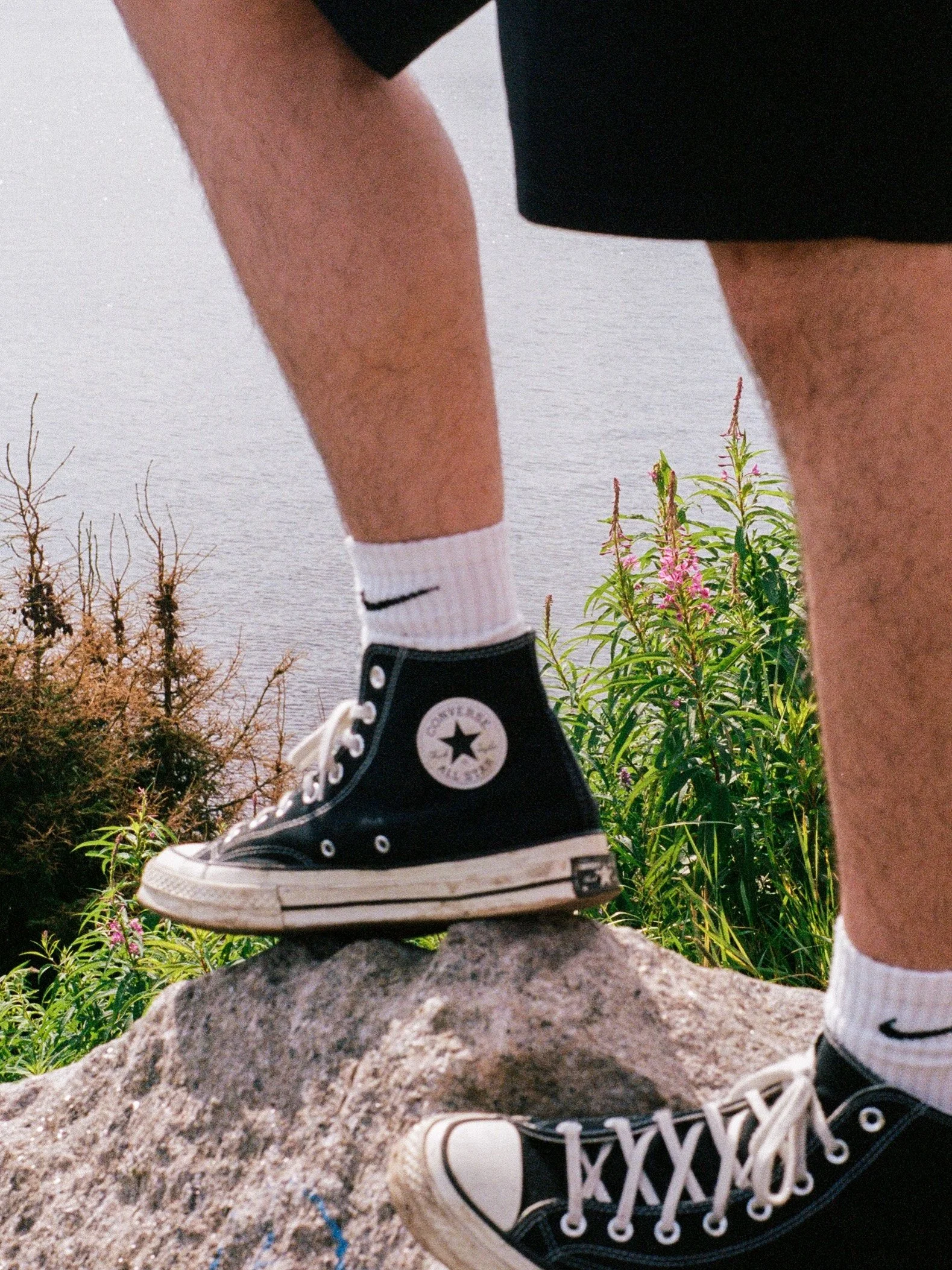 Une personne portant des chaussures Converse noires, des chaussettes blanches avec un logo Nike, et un short noir, se tient sur un rocher à côté d'une rivière ou d'un lac avec des plantes autour.