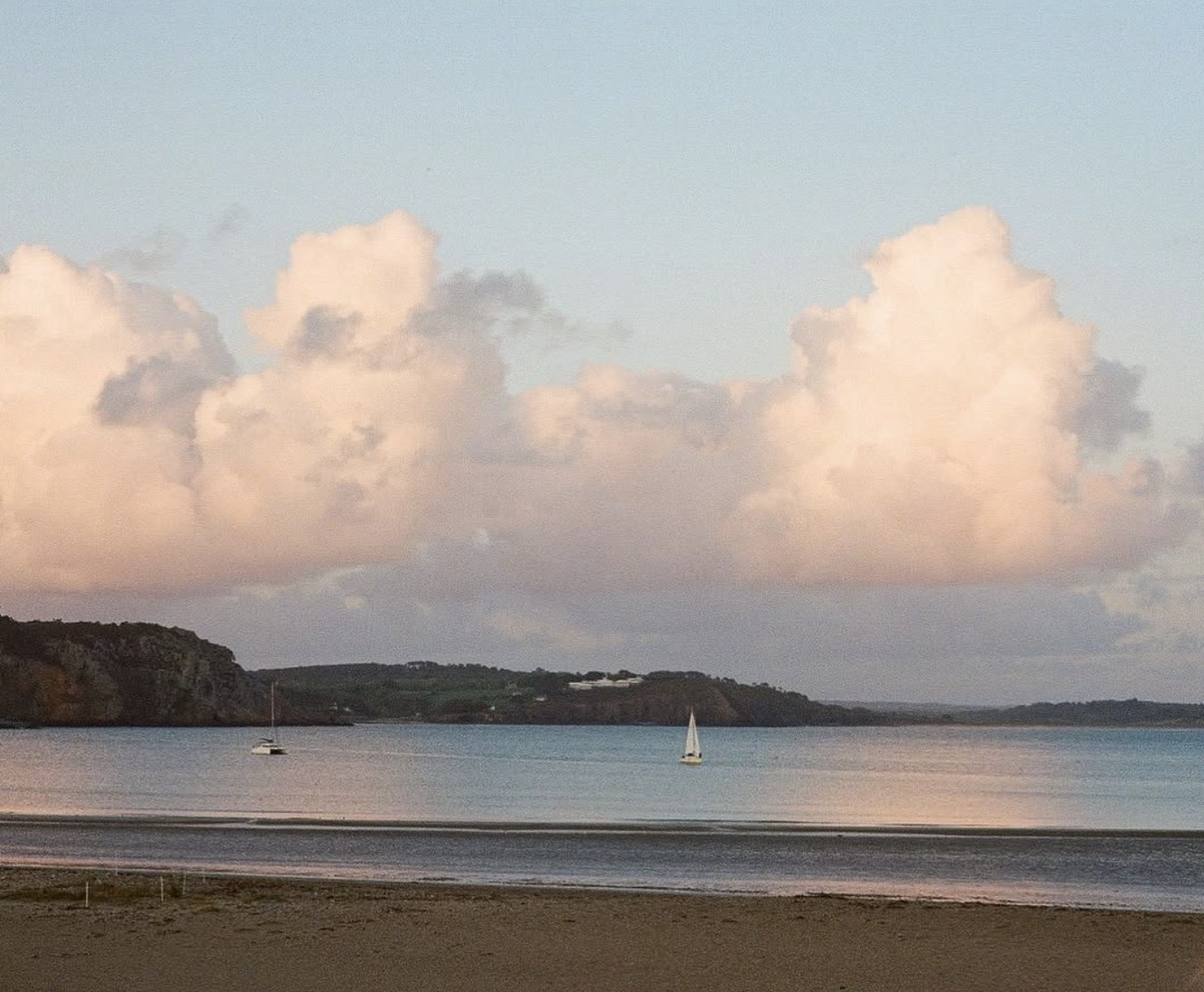 Ciel nuageux avec deux voiliers sur un plan d'eau calme, côte rocheuse en arrière-plan au coucher du soleil.