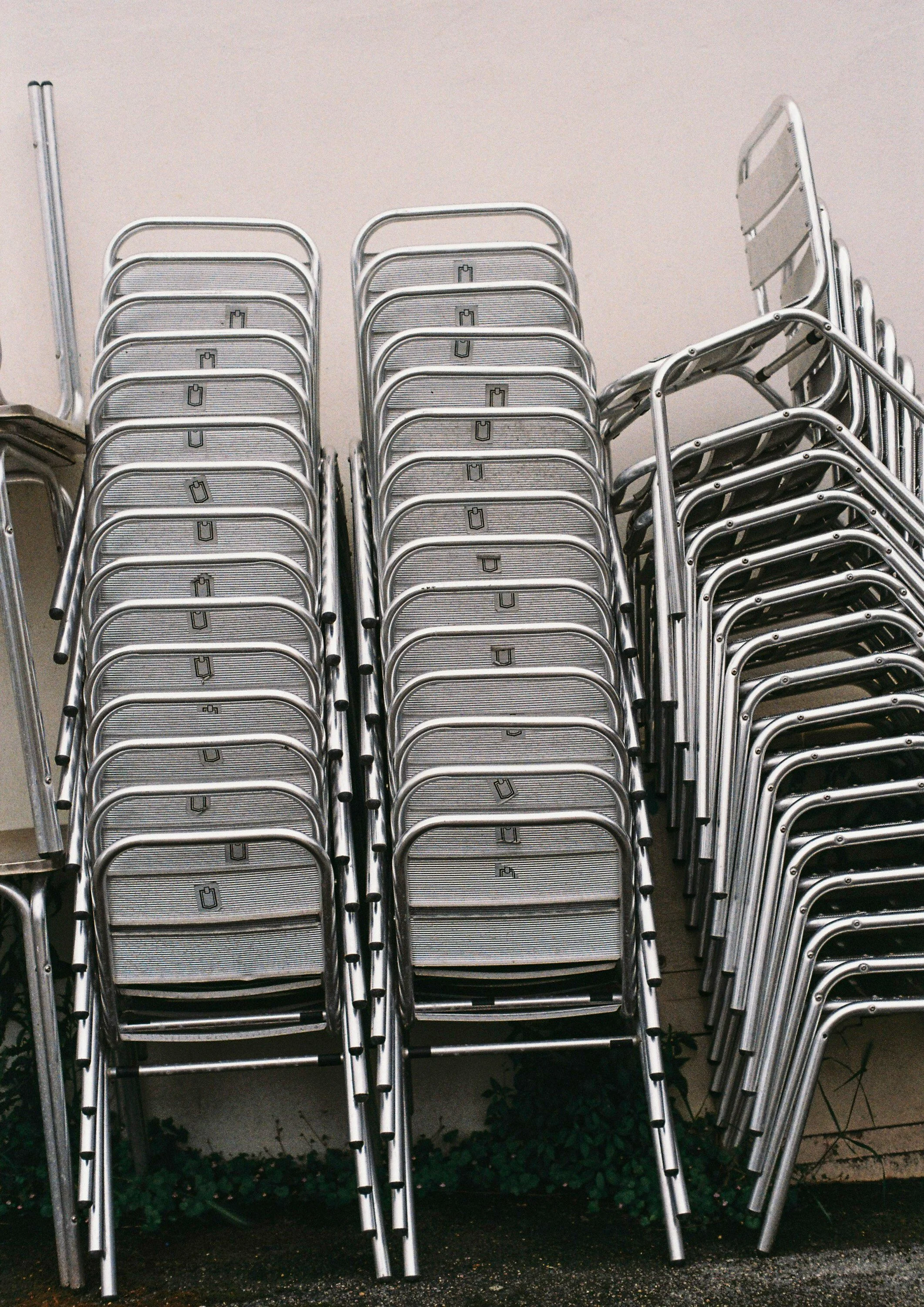 Plusieurs chaises métalliques empilées et des chaises pliantes posées contre un mur beige, avec un petit espace végétal en dessous.