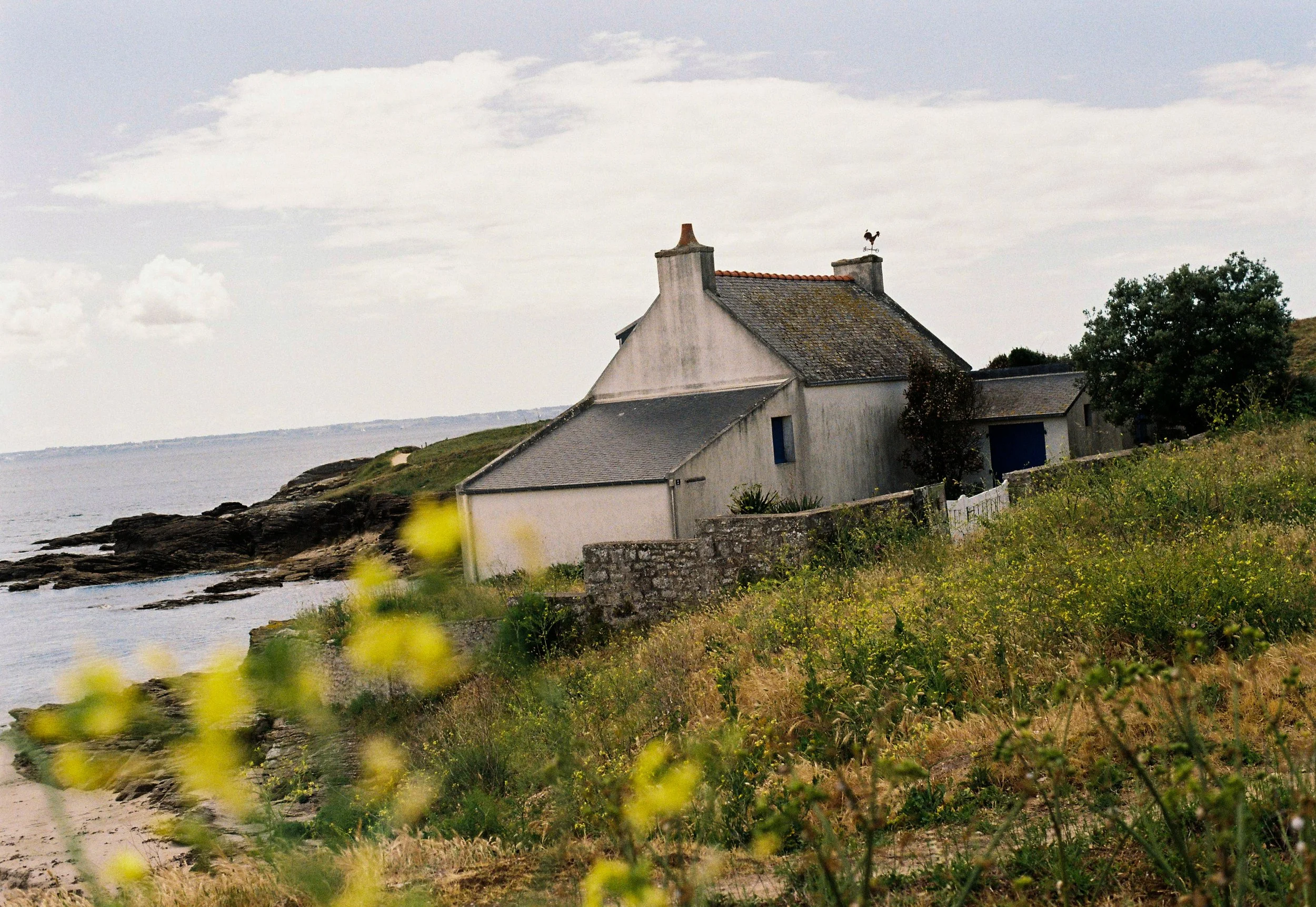 Maisons blanches avec cheminée sur une colline au bord de la mer, entourées de végétation verte et de rochers, sous un ciel nuageux.