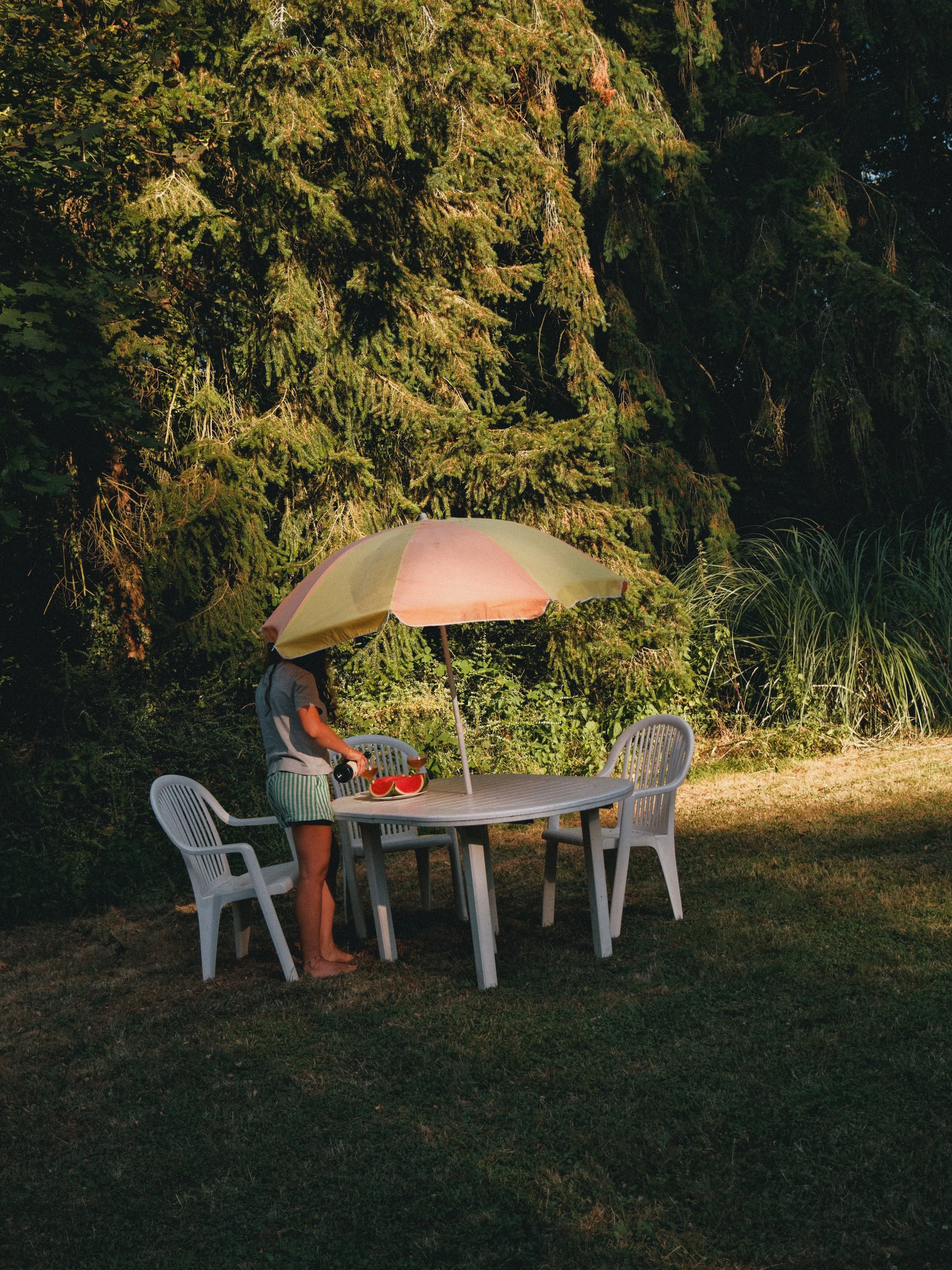 Une personne se tient à côté d'une table en plastique blanche sous un grand parasol jaune et rose dans un jardin entouré d'arbres et d'herbes vertes, en soirée.
