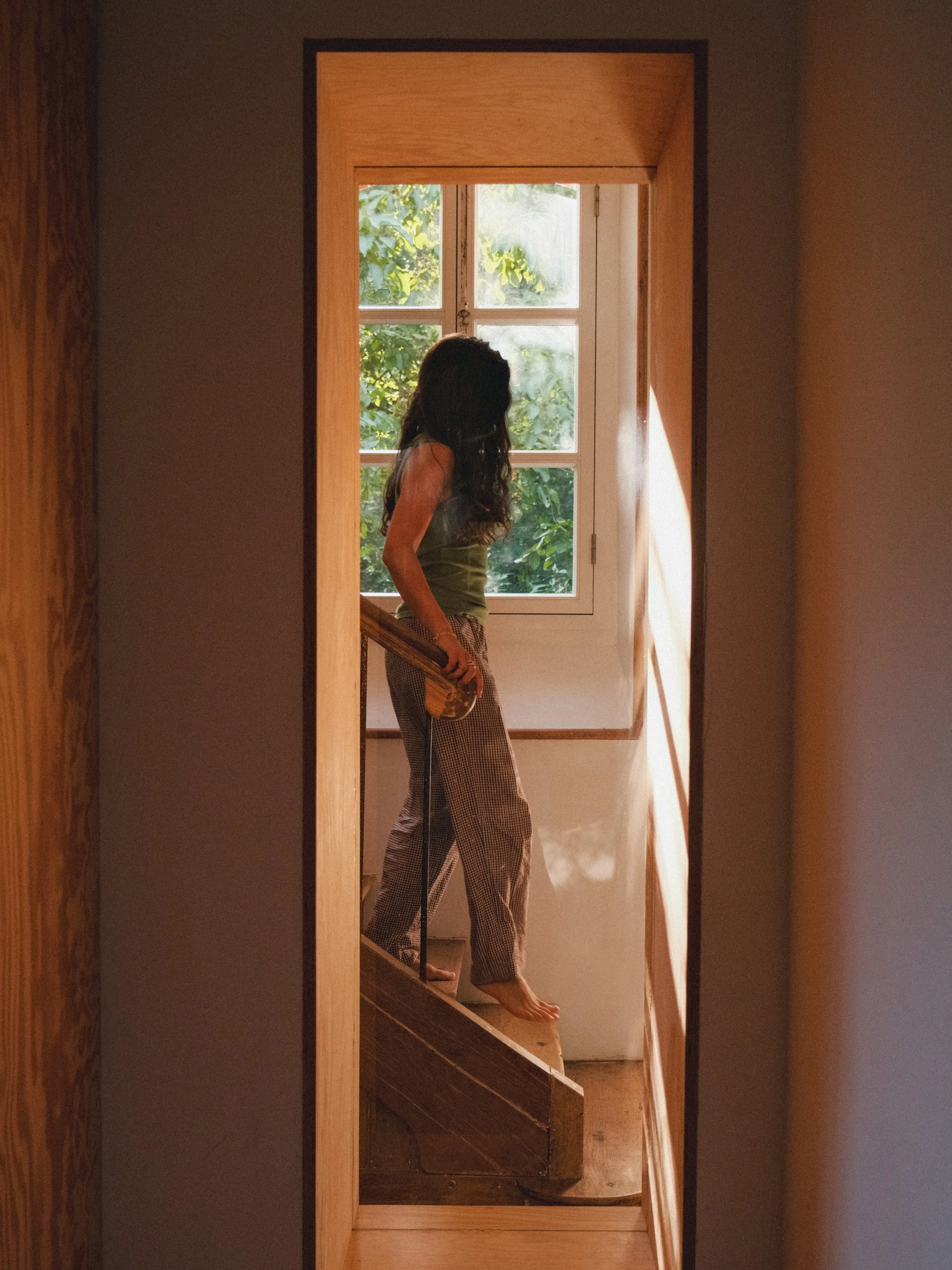 Femme avec de longs cheveux bruns, portant un haut sans manches vert et un pantalon à carreaux, debout à l'intérieur, près d'une fenêtre avec des volets en bois, en train de descendre un escalier en bois.