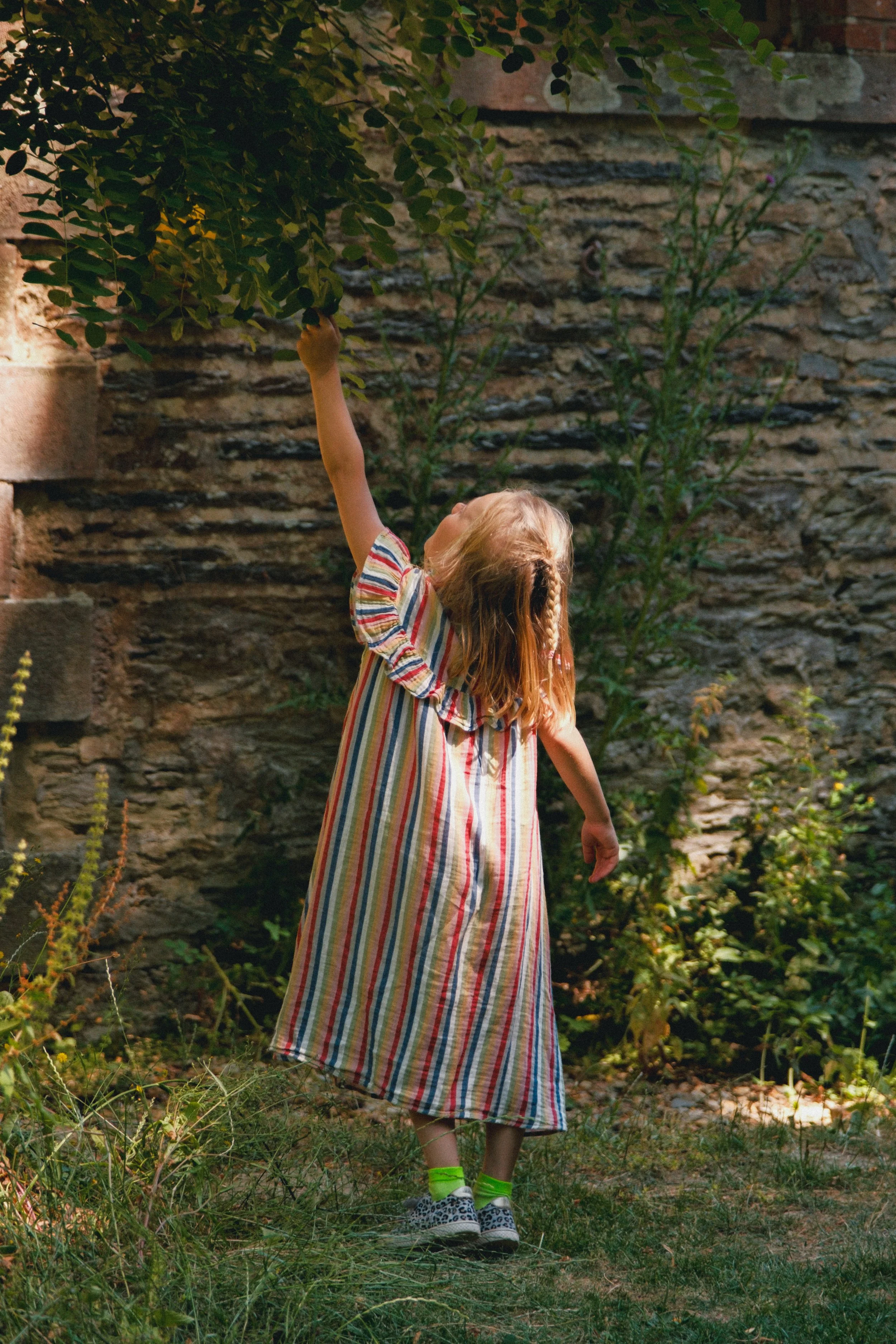 Une jeune fille aux cheveux longs et blonds portant une robe à rayures, des tennis à motifs léopard, et des chaussettes vertes, touche les feuilles d'un arbre dans un jardin avec un mur en pierre en arrière-plan.