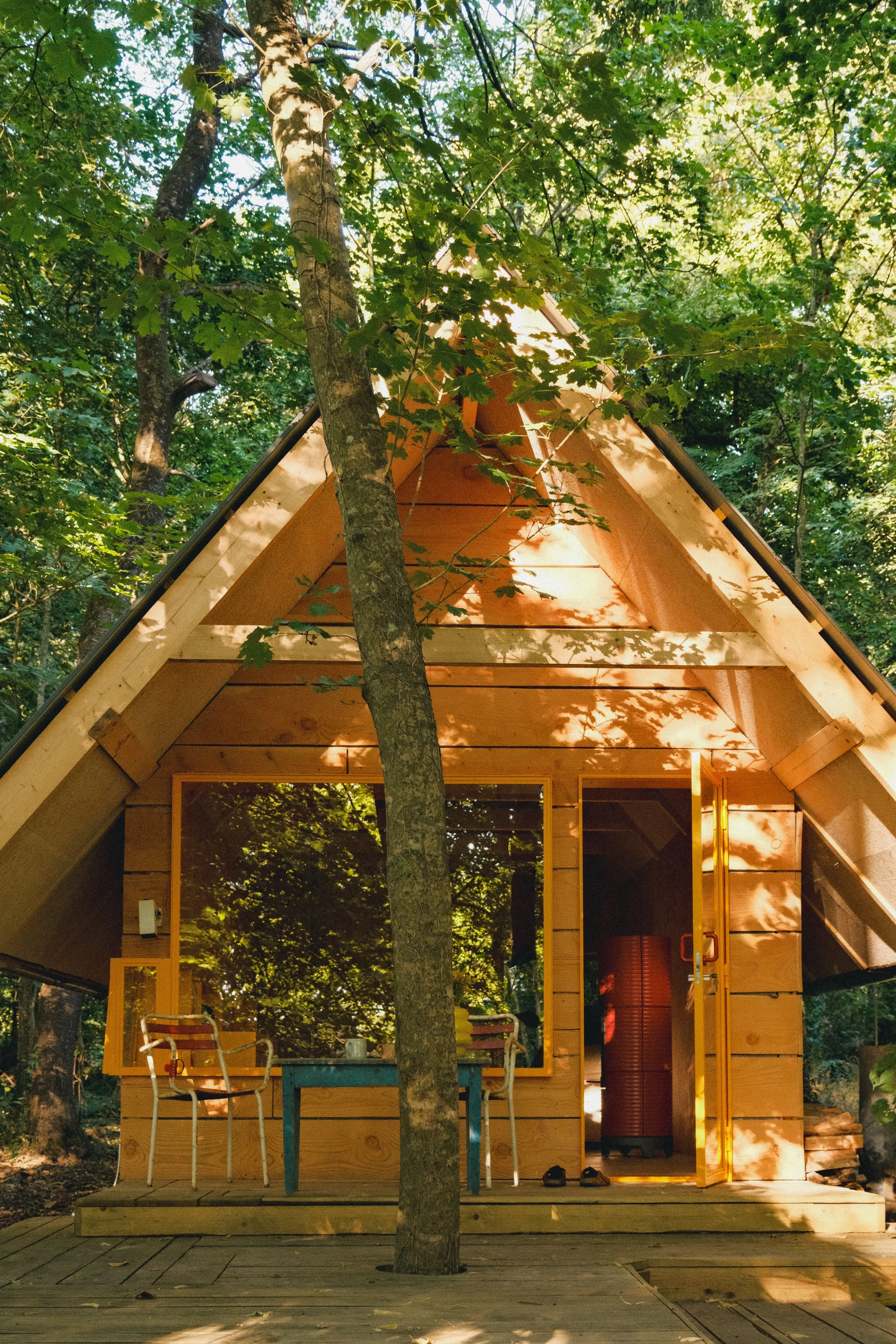 Une cabane en bois dans une forêt, entourée d'arbres et de feuilles, avec un espace extérieur comprenant une table, des chaises, et une grande bouteille rouge à l'intérieur.