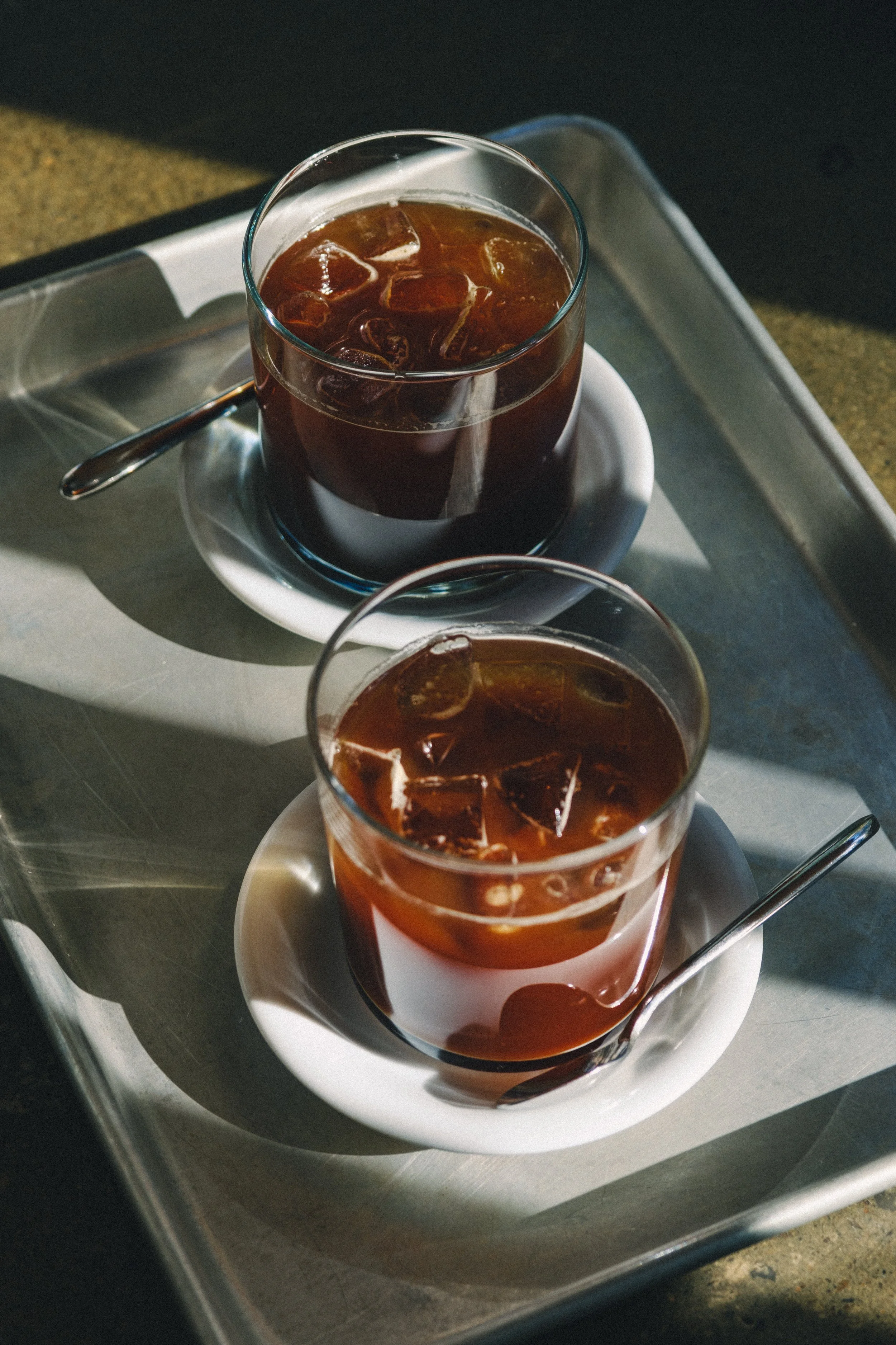 Deux verres de café glacé sur une plateau en métal, la boisson est sombre avec des glaçons et une cuillère dans chaque verre.