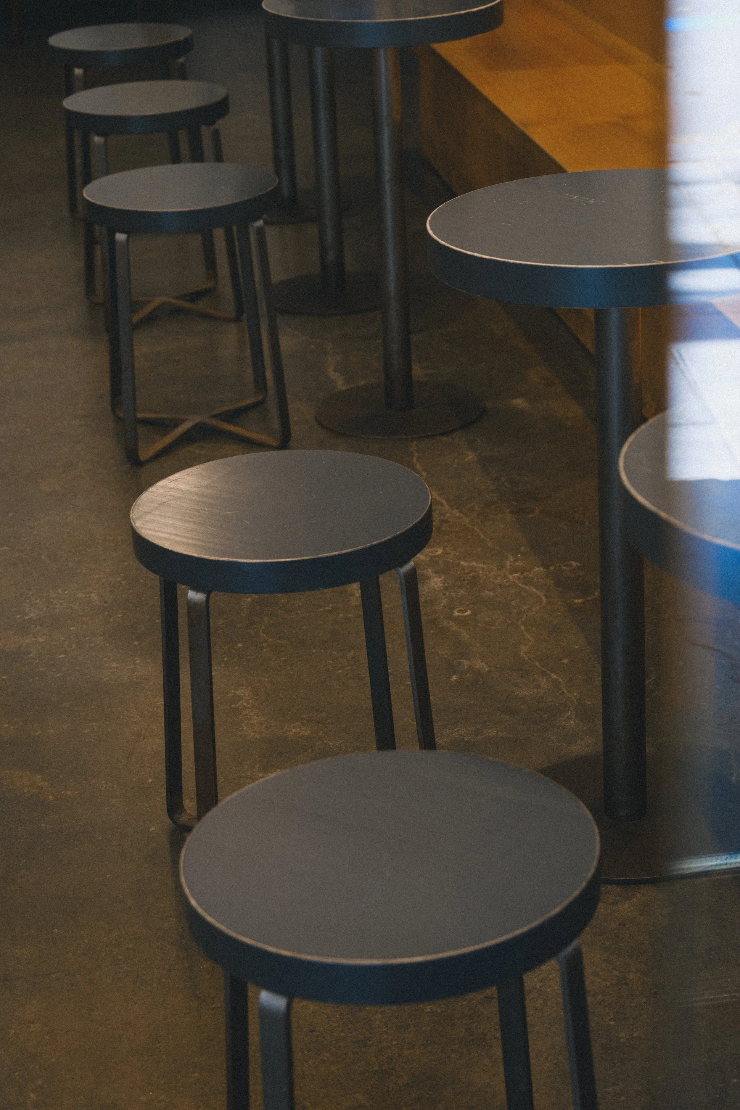 Série de petites tables rondes noires avec des tabourets assortis dans un restaurant ou un café, disposés près d'une banquette en bois.