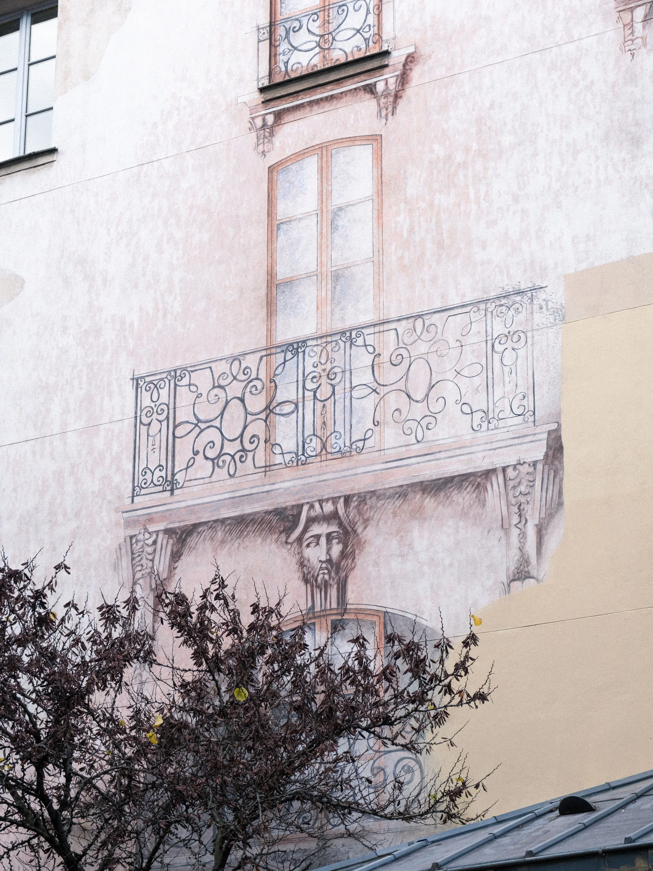 Fresque murale représentant une fenêtre de balcon avec un décor en fer forgé, de style ancien, et un visage d'homme peint en dessous, avec un arbre feuillu en premier plan.