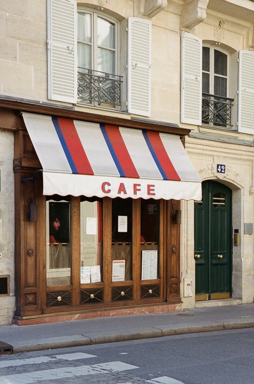 Façade d'un café avec une enseigne en tissu rayé rouge, blanc et bleu, portant le mot « CAFE ». fenêtres à volets blancs et une porte verte, numéro 42.