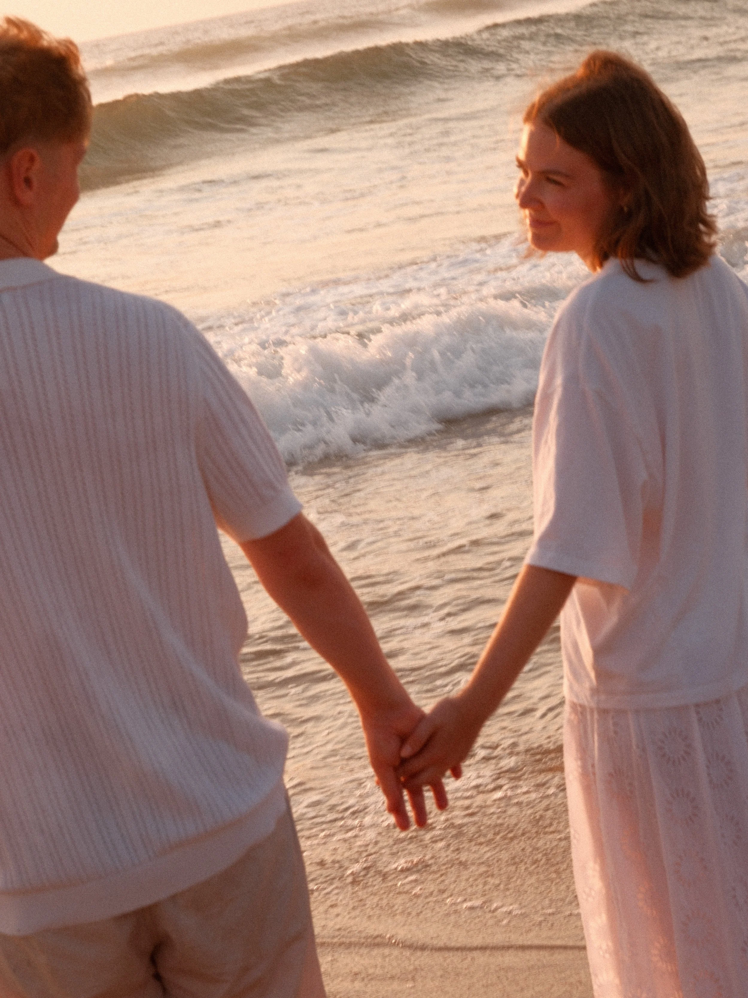 Deux personnes se tenant la main sur la plage au coucher du soleil, vêtues de tenues légères.