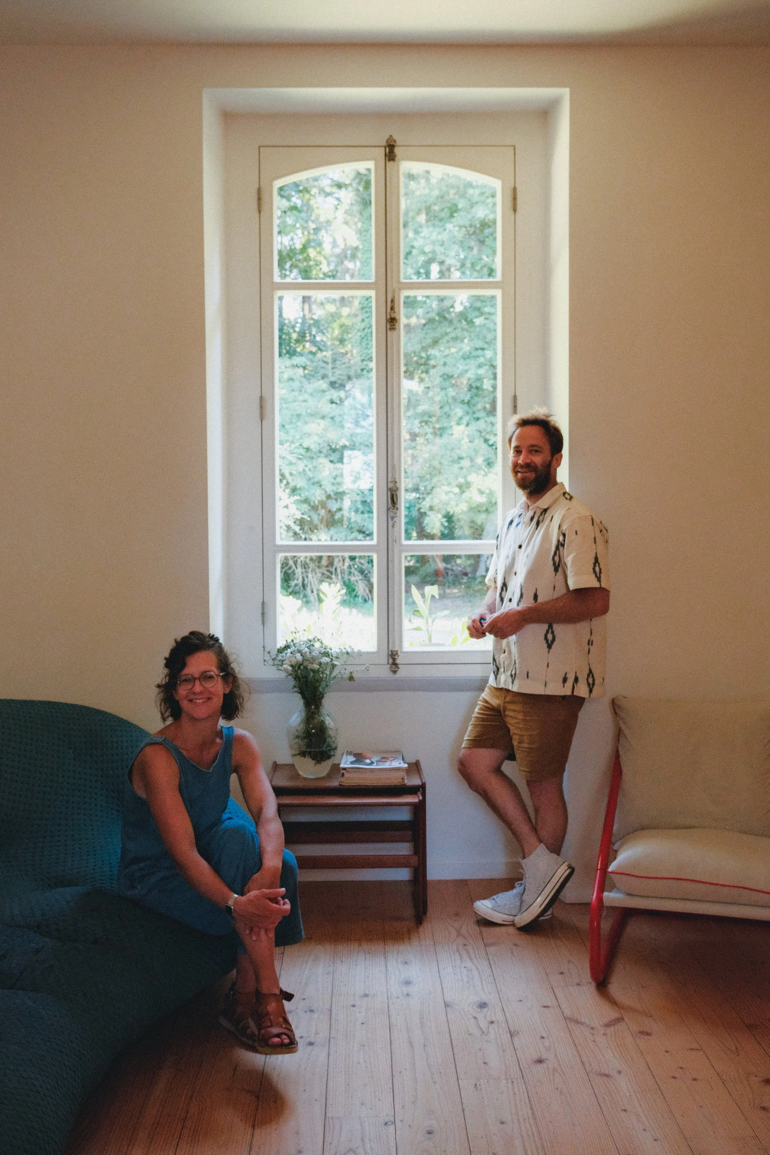 Deux personnes, une femme assise sur un canapé et un homme debout près d'une fenêtre, dans une pièce lumineuse avec un sol en bois, une petite table avec un vase de fleurs, une pile de livres, et un coussin beige.