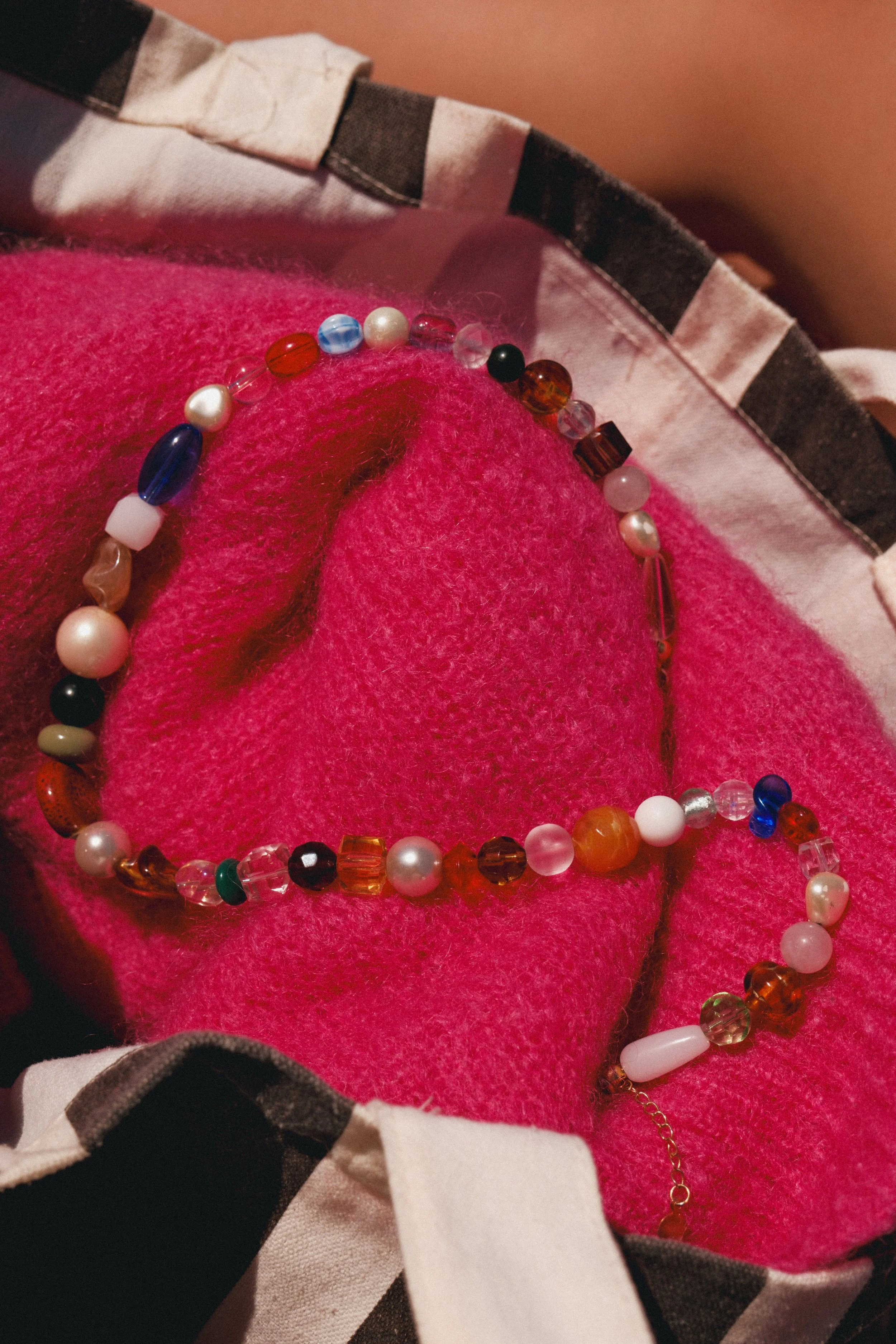 Collier de perles multicolores posé sur un tissu rose. Présence d'une chaîne dorée en partie visible.