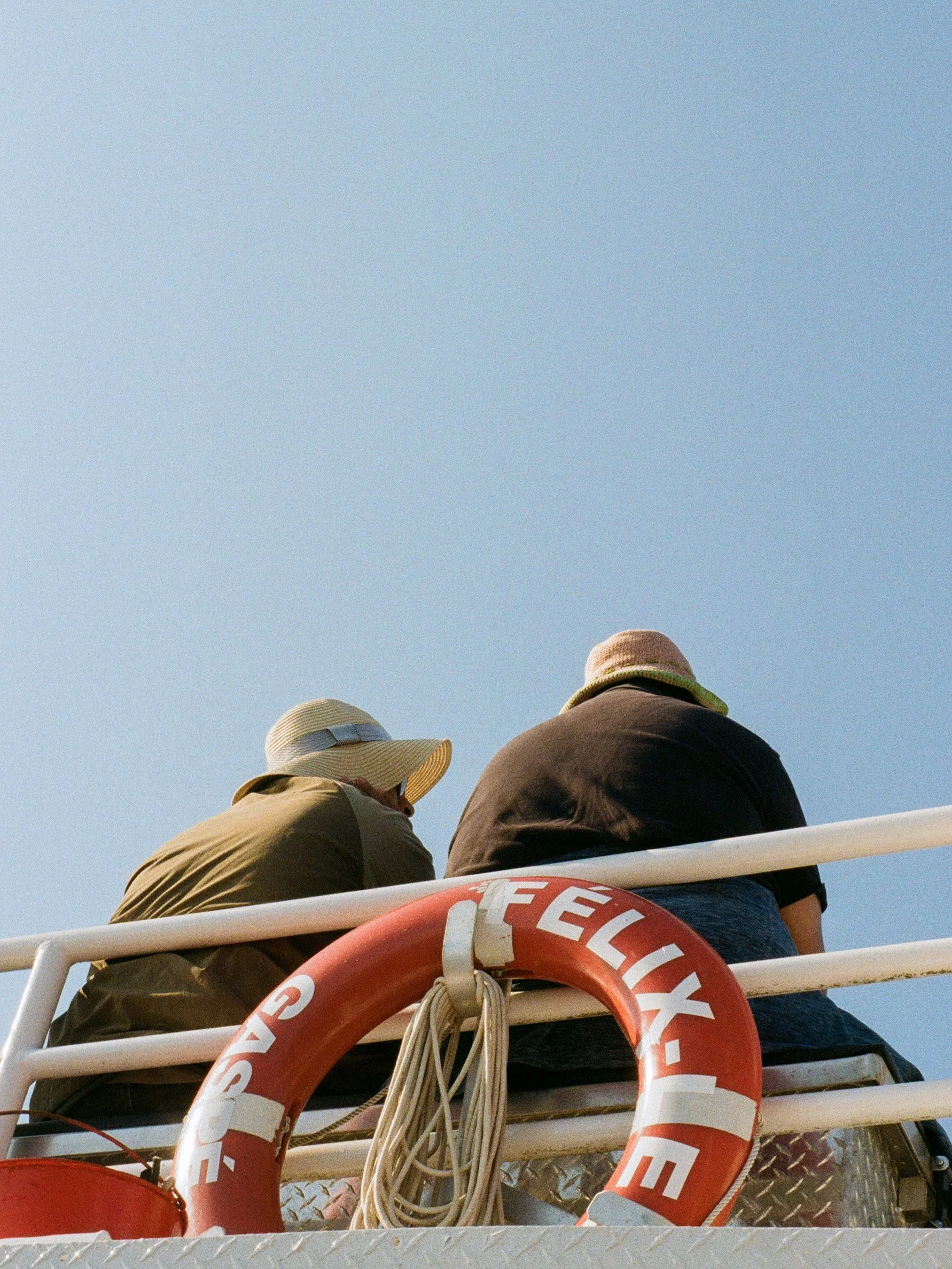 Deux personnes portant des chapeaux, assises à l'arrière d'un véhicule ou embarcation, avec un cercle de vie de sauvetage orange marqué 'RESCUE' et des cordes, sous un ciel clair.