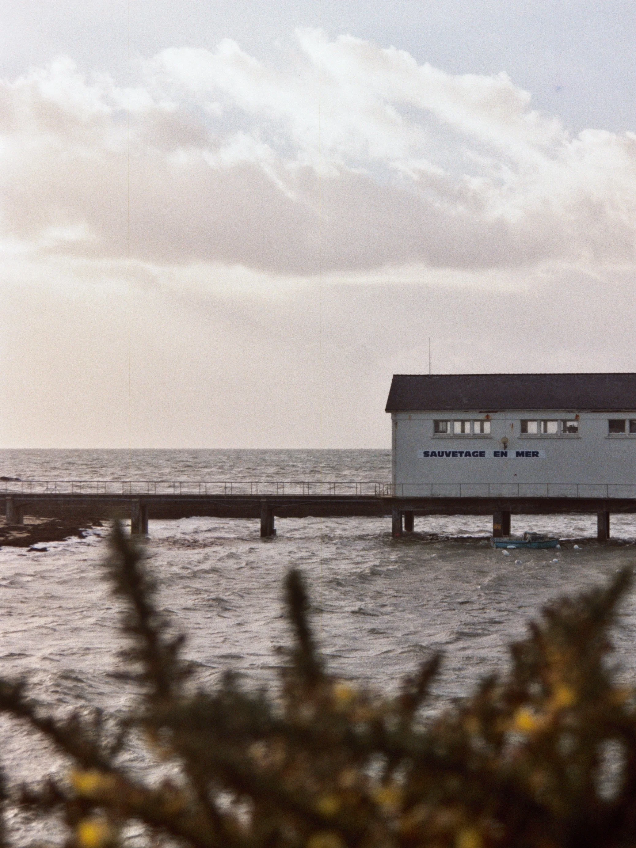 Une maison en bord de mer avec une pancarte indiquant "Sauvetage en mer" vue depuis la plage, avec des nuages et une mer agitée en arrière-plan.