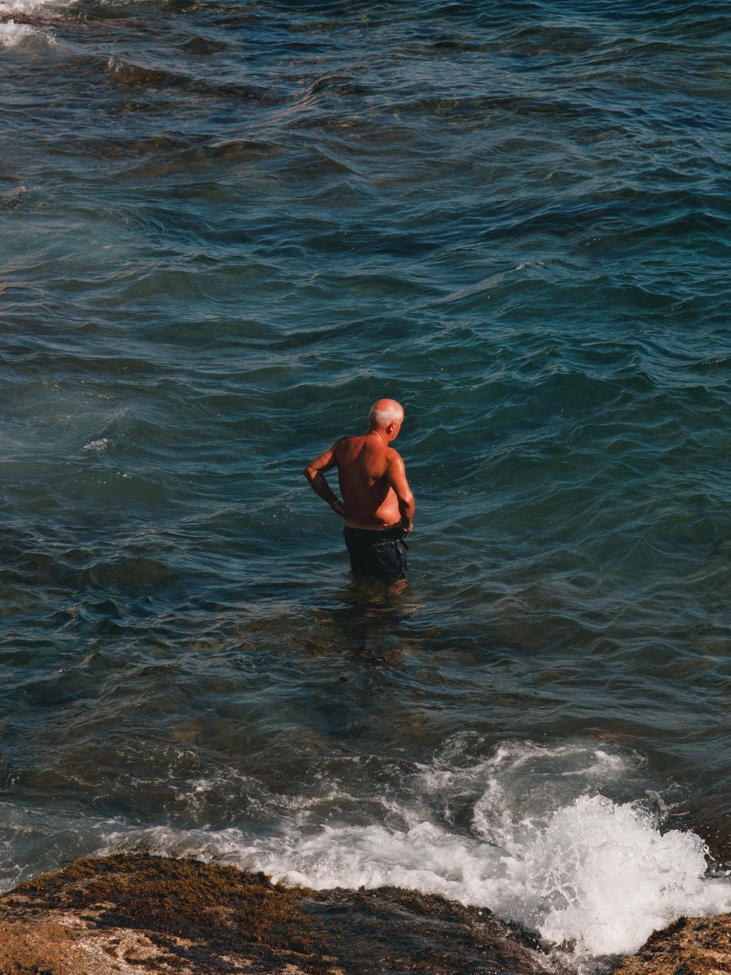 Un homme vieux, chauve, en maillot de bain noir, marche dans la mer près des rochers.