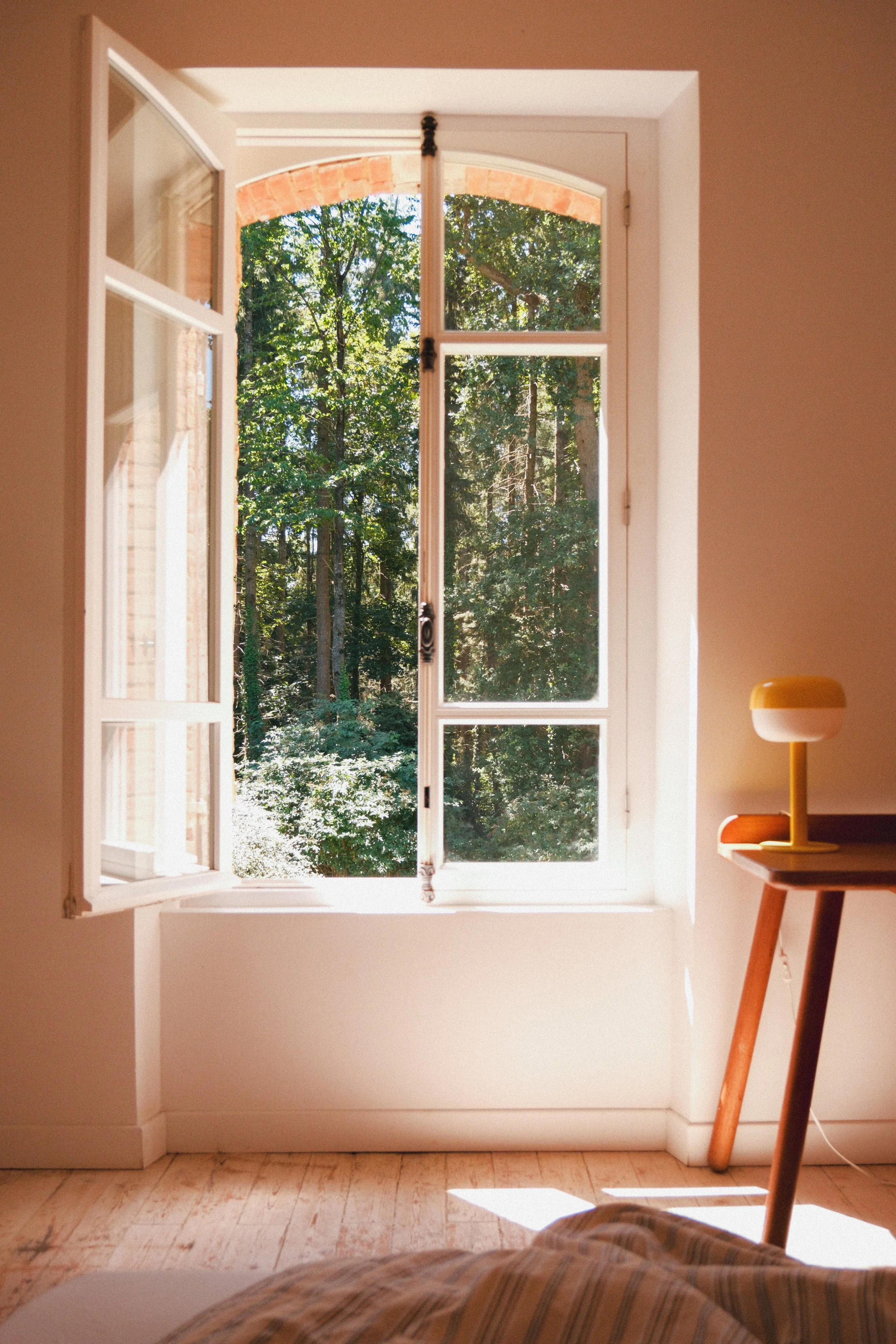 Vue d'une fenêtre ouverte donnant sur une forêt avec des arbres verts, à l'intérieur d'une pièce avec un sol en bois clair, un lampadaire au côté droit et un lit avec une couverture en tissu à rayures dans le premier plan.