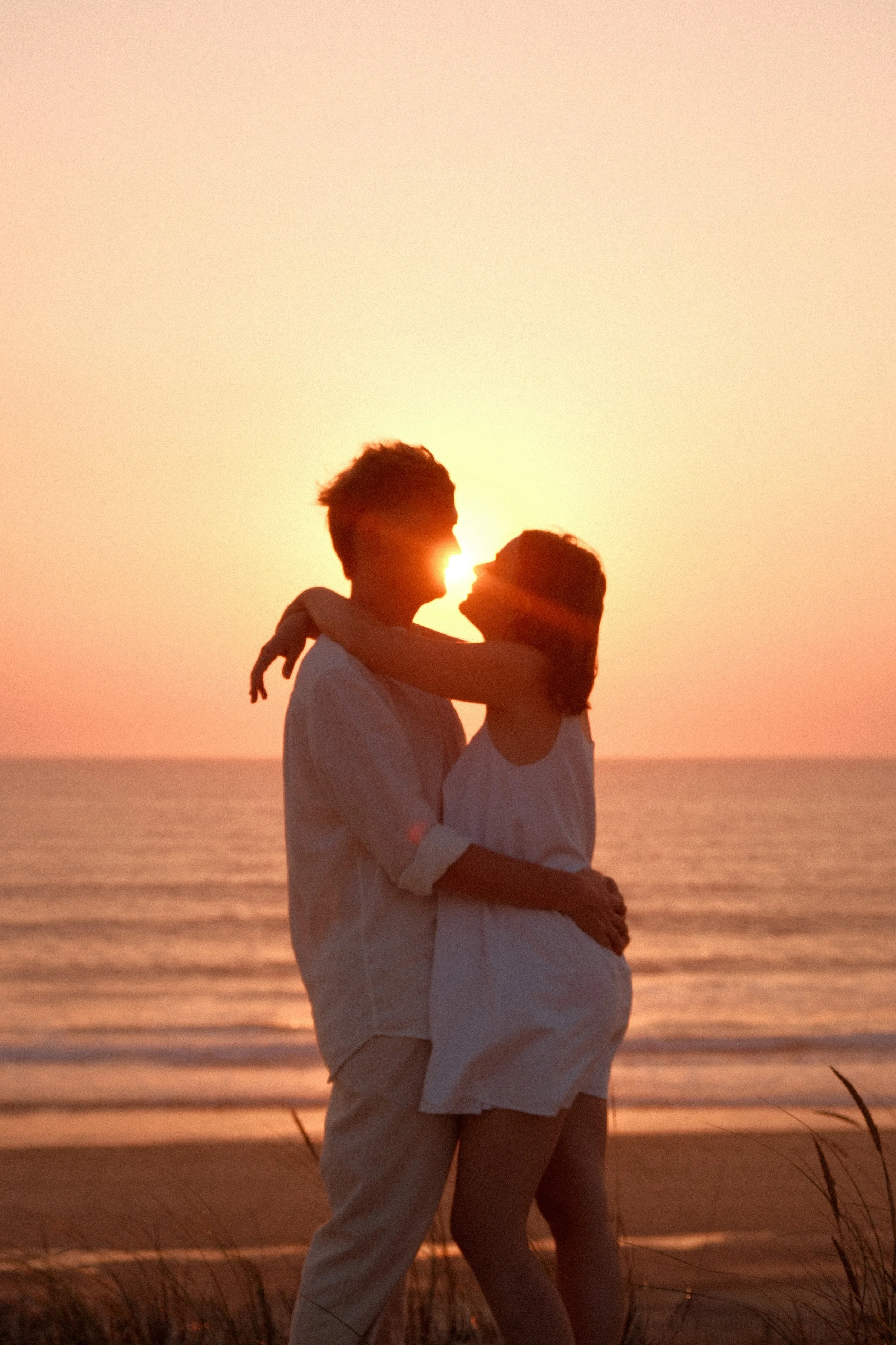 Un couple souriant et s'étreignant sur la plage au coucher du soleil, avec la mer en arrière-plan.