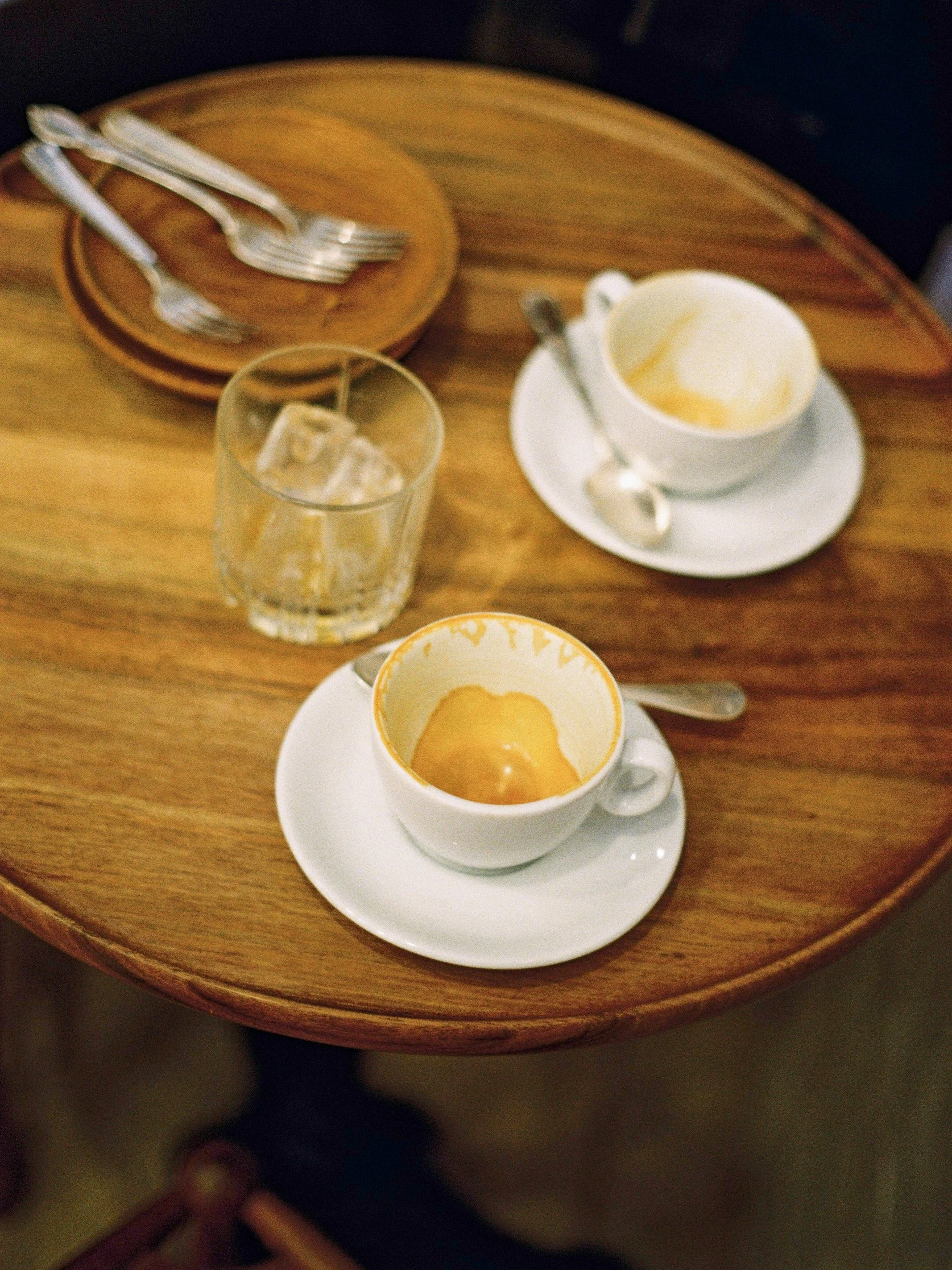 Tasse de café vide avec trace de café dans une assiette blanche, à côté d'un verre avec glaçons et une cuillère, sur une table en bois.