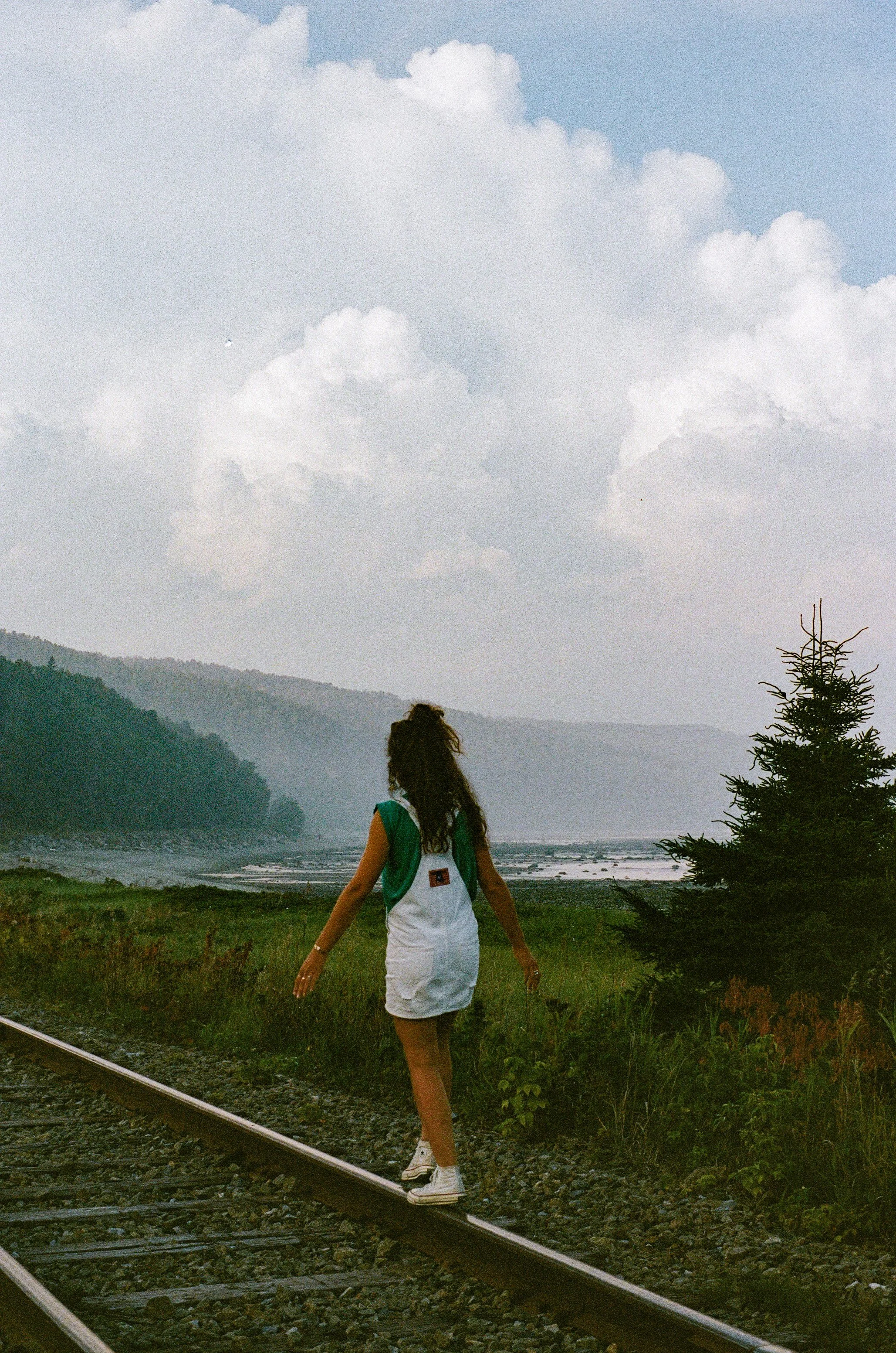 Une femme marche près de voies ferrées dans un paysage naturel avec des montagnes au loin et un ciel nuageux.