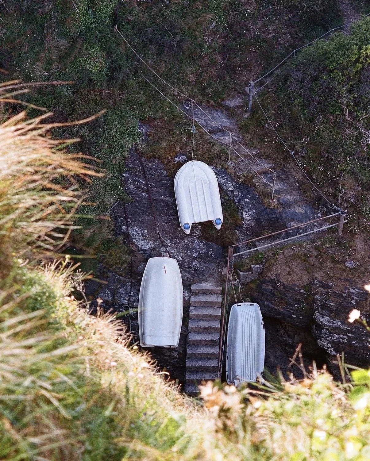 Trois canots blancs suspendus à une falaise avec escaliers en pierre, entourés de végétation.