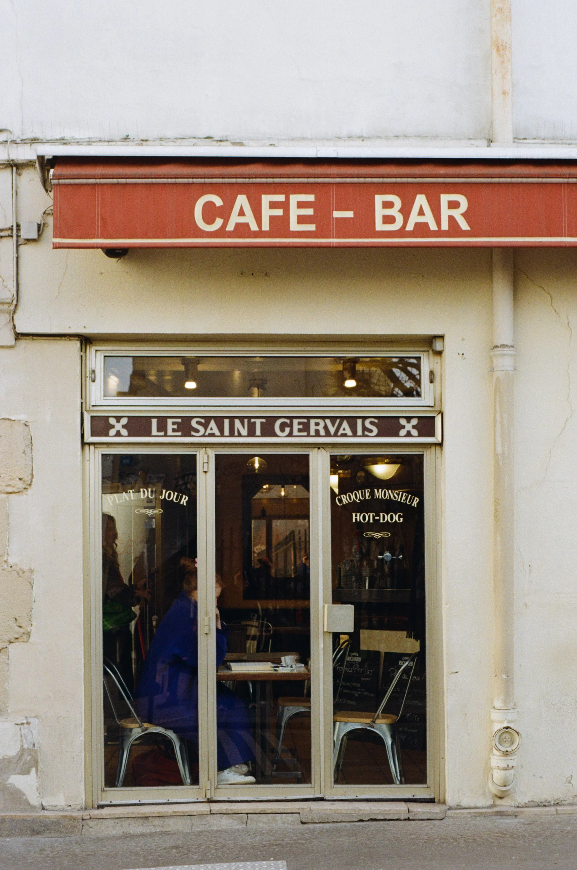 Façade d'un café-restaurant avec vitrine et porte en verre, intérieur visible avec des clients et des panneaux de menu.