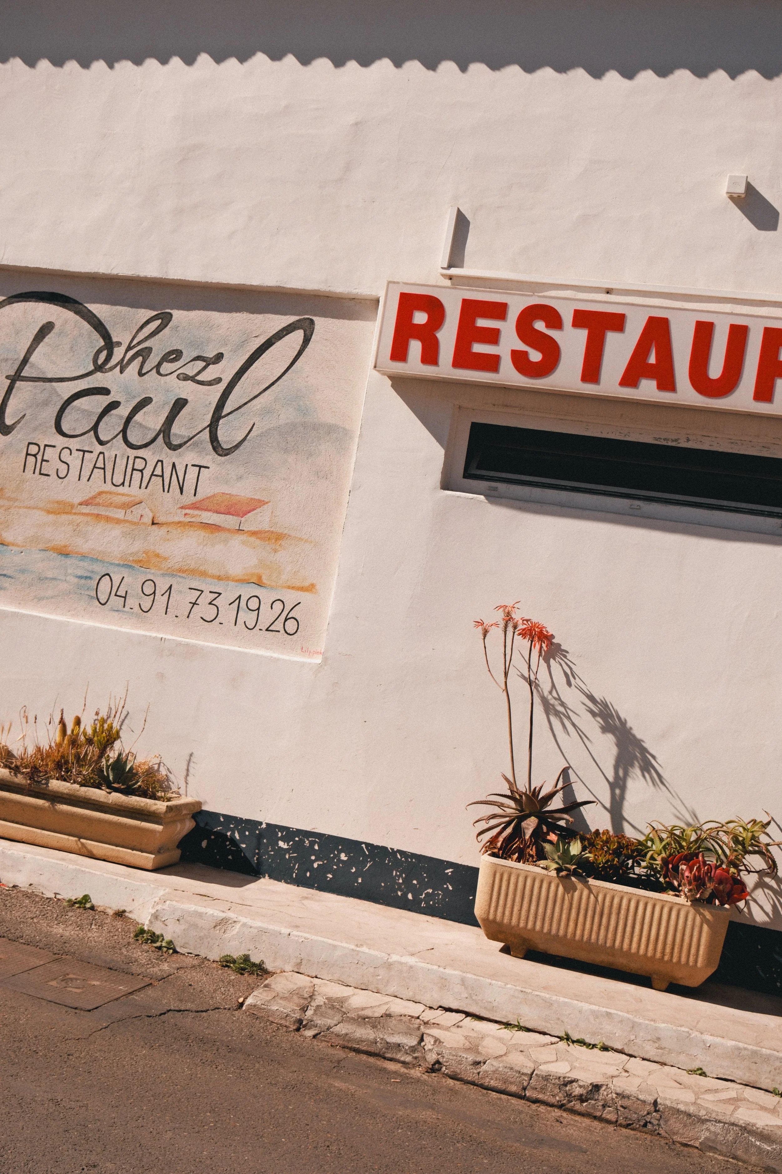 Vue d'une façade de restaurant avec un panneau rouge et blanc 'RESTAURANT' et un autre panneau avec le nom 'Chez l'Aul', des plantes en pot et un trottoir en pavés.