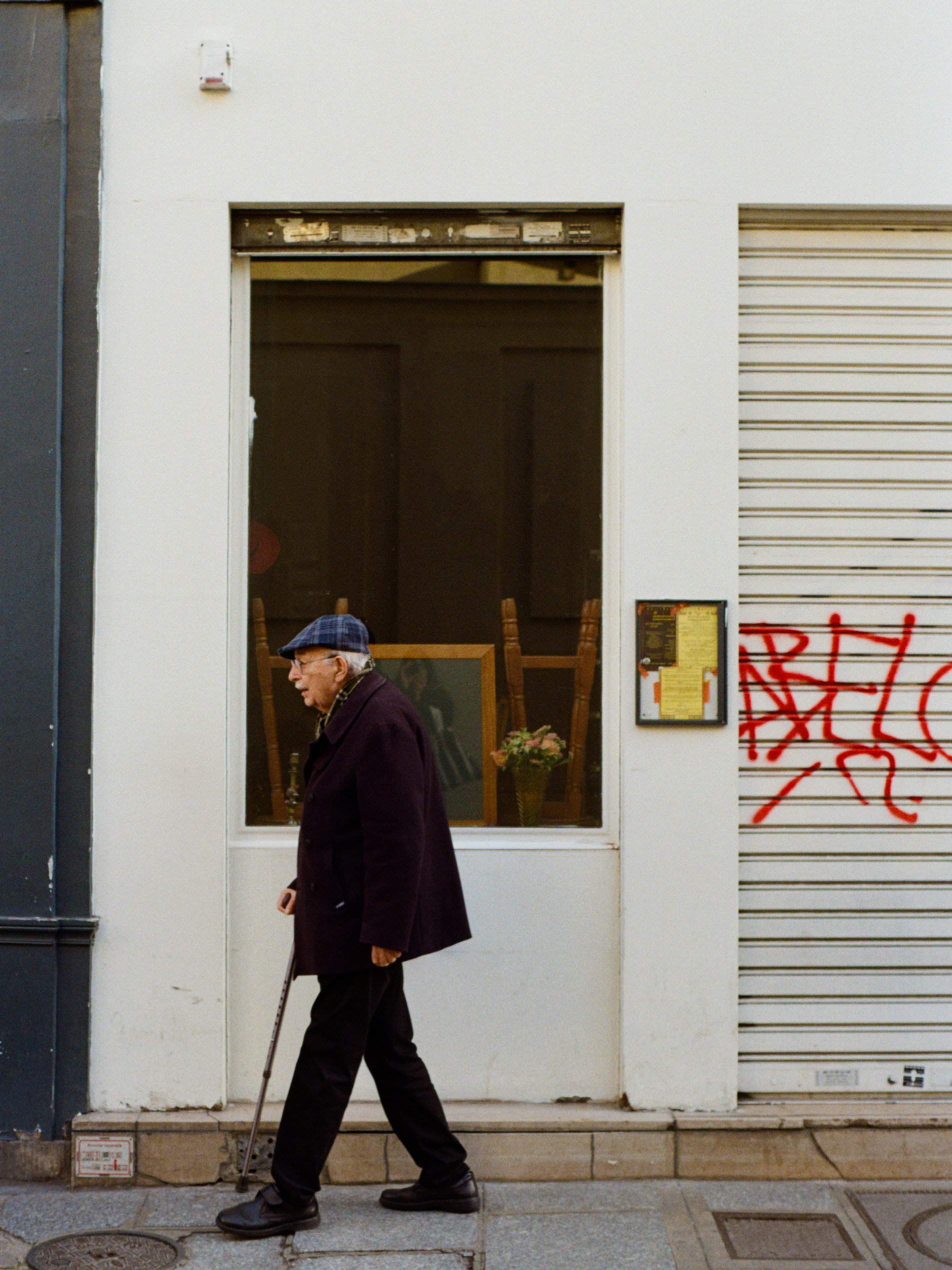 Un homme âgé avec un bâton marche devant un bâtiment blanc avec une fenêtre et une porte à volets roulants gris, un panneau d'informations à côté de la porte, et un graffiti rouge sur le volet roulant.