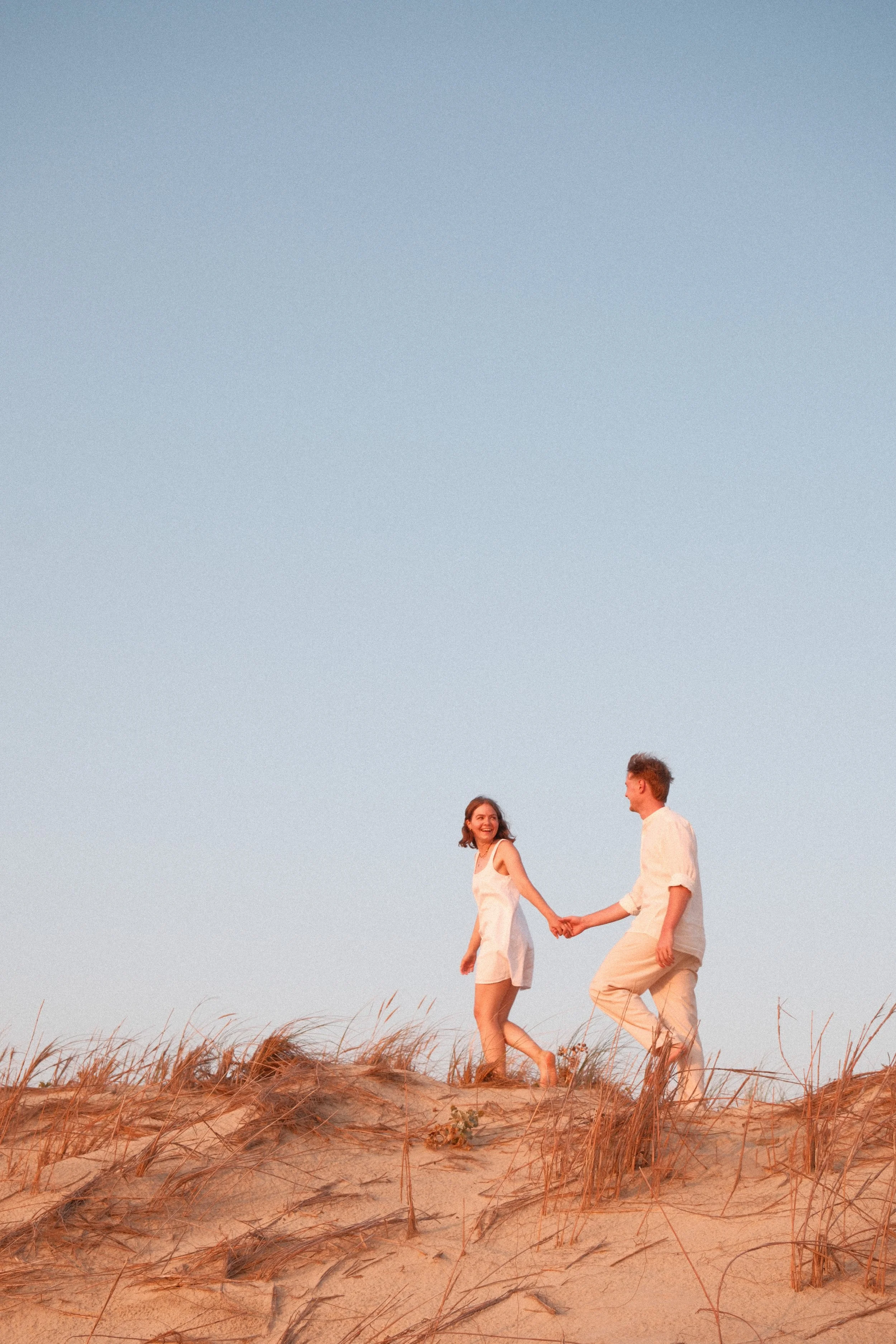 Un couple marchant main dans la main sur une plage de sable avec des herbes hautes, sous un ciel clair.