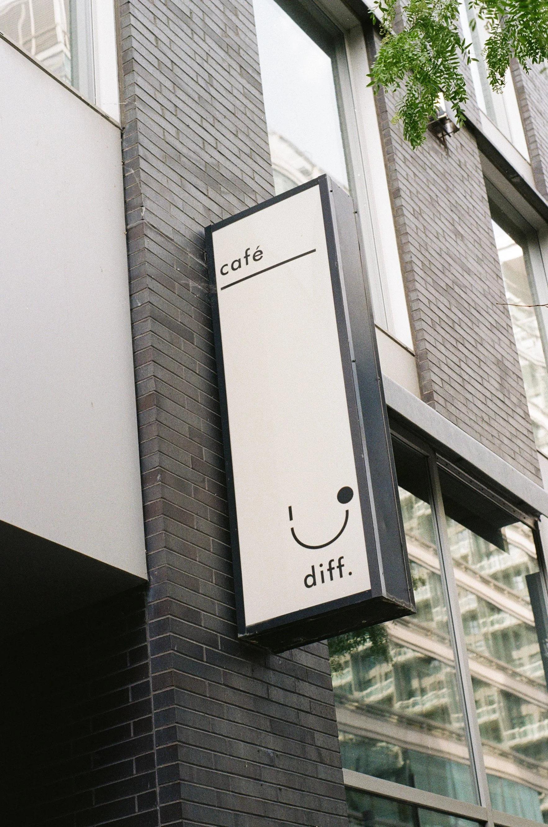 Panneau de signalisation extérieure pour un café, avec une illustration d'un sourire stylisé et le texte 'café diff.' sur un fond blanc, fixé à un mur en briques noires