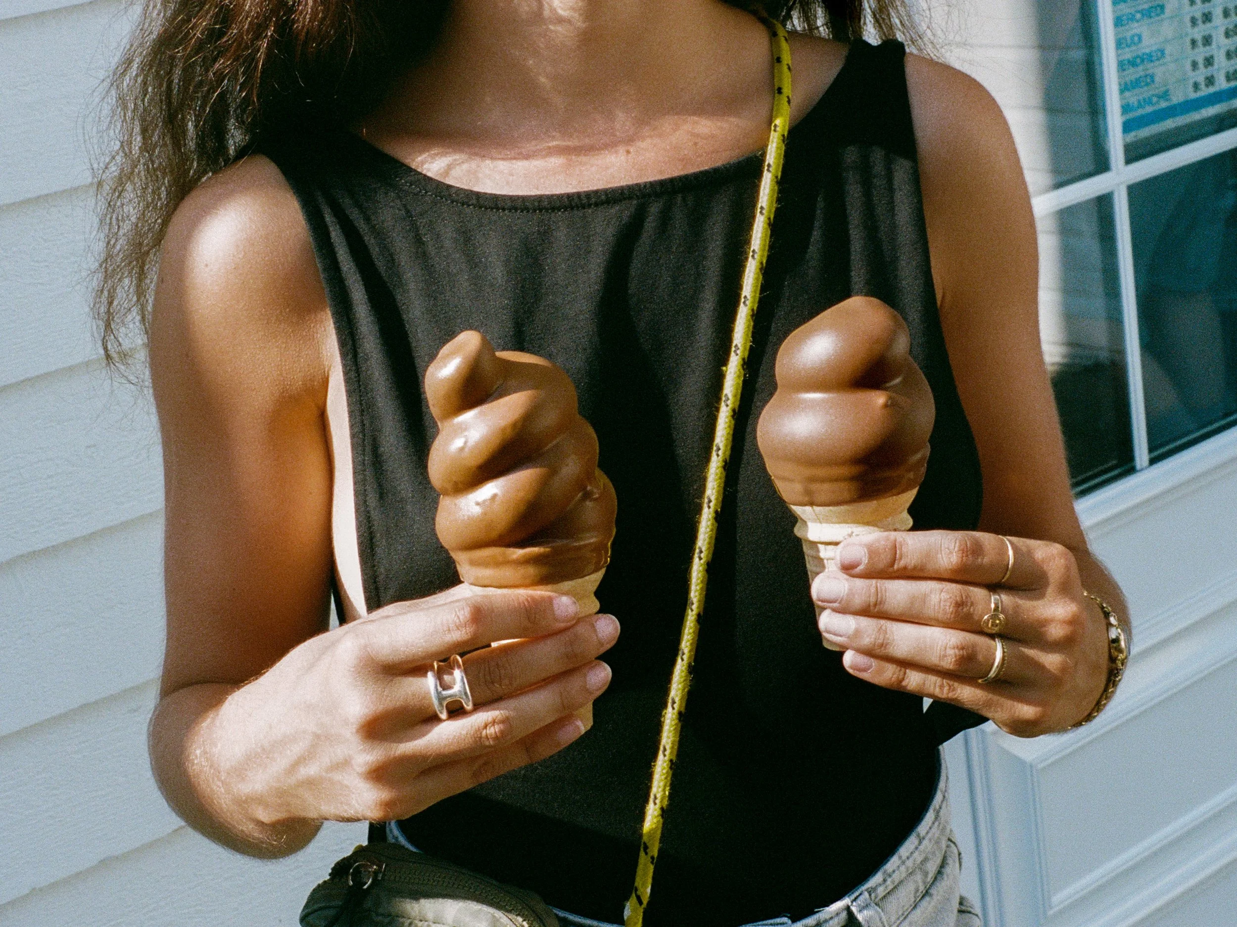 Une personne portant un haut noir tissé en dentelle, tenant deux glaces au chocolat. Elle porte plusieurs bagues dorées et un bracelet, et a une règle jaune enroulée autour du cou. La scène se déroule devant une façade en bardeaux blancs et une fenêt