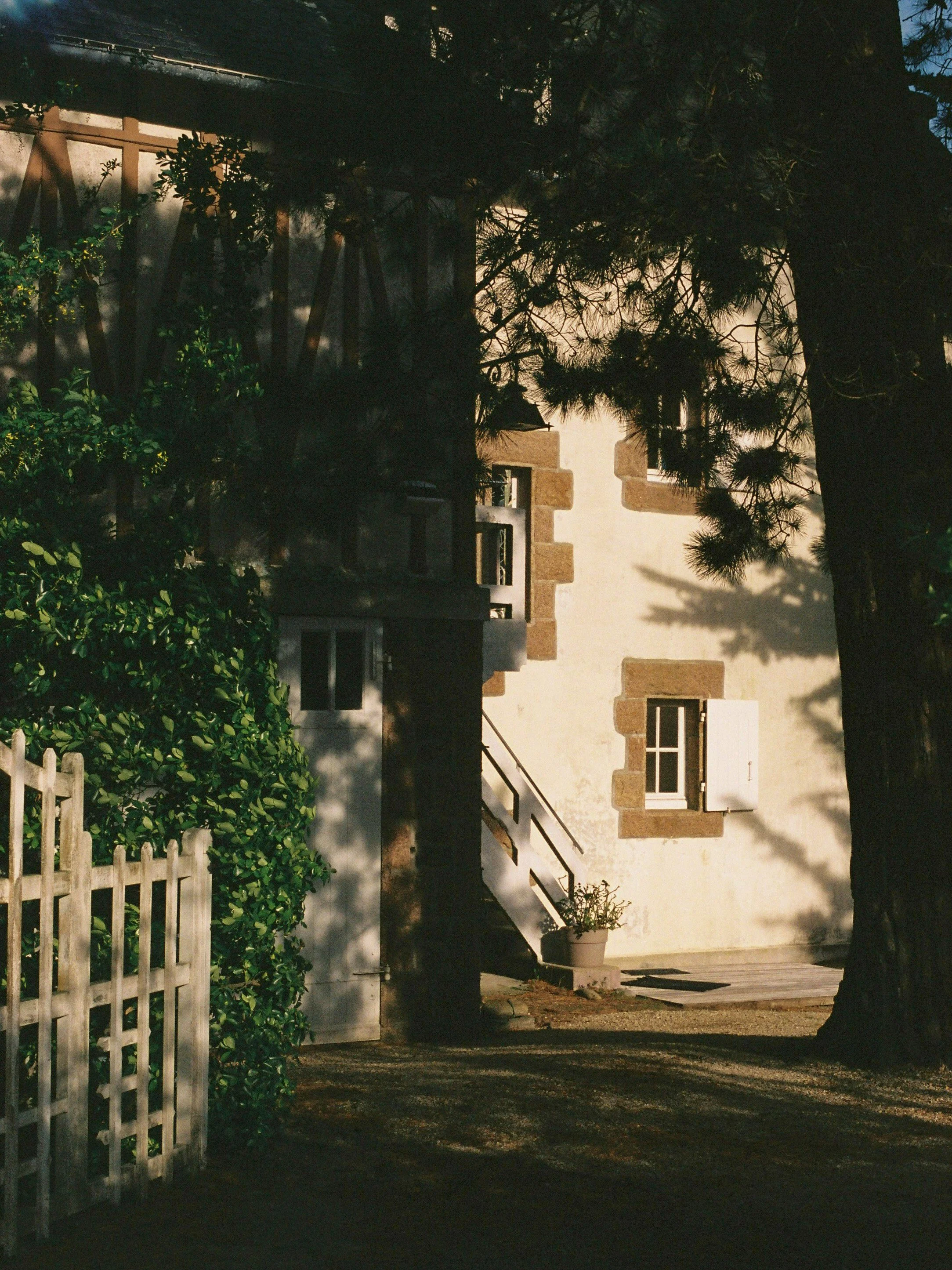 Une maison avec de petites fenêtres à la façade blanche encadrée de pierre, entourée d'arbres et d'une clôture en bois, en plein air avec un sol en terre et un pot de fleurs blanc.