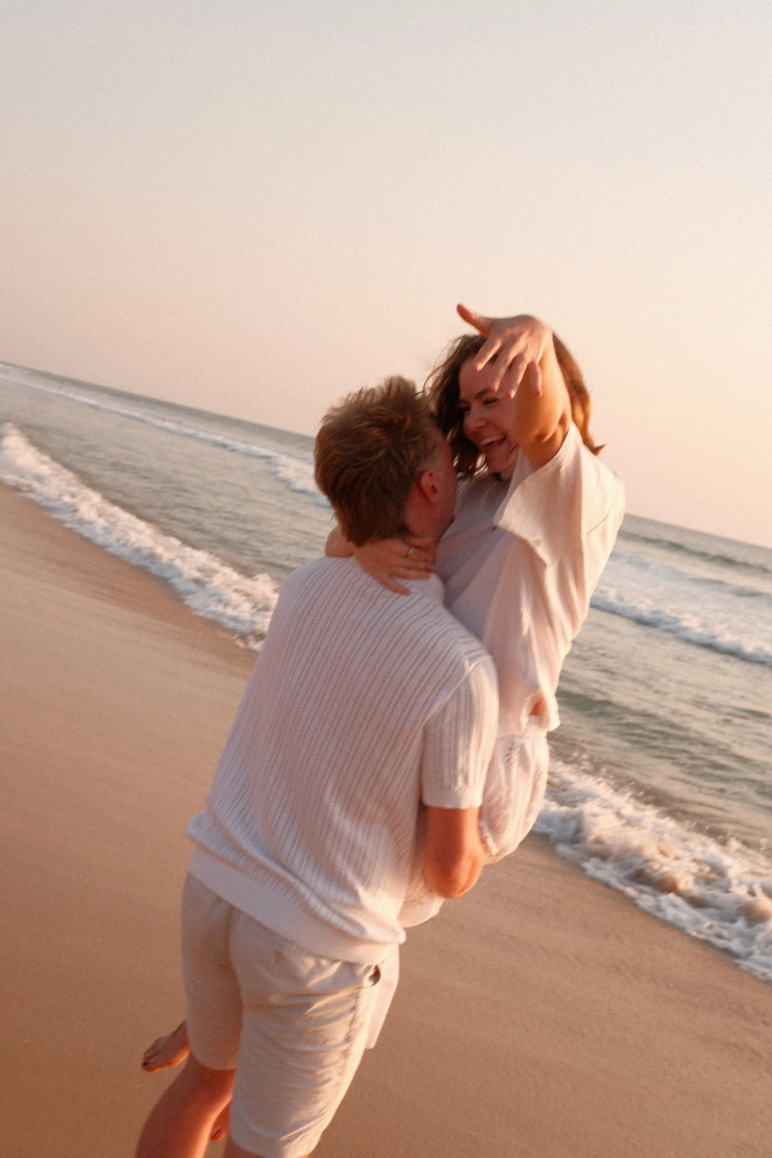 Un couple au bord de la mer, l'homme soulève la femme dans ses bras, tous deux souriants, la plage au coucher du soleil en arrière-plan.