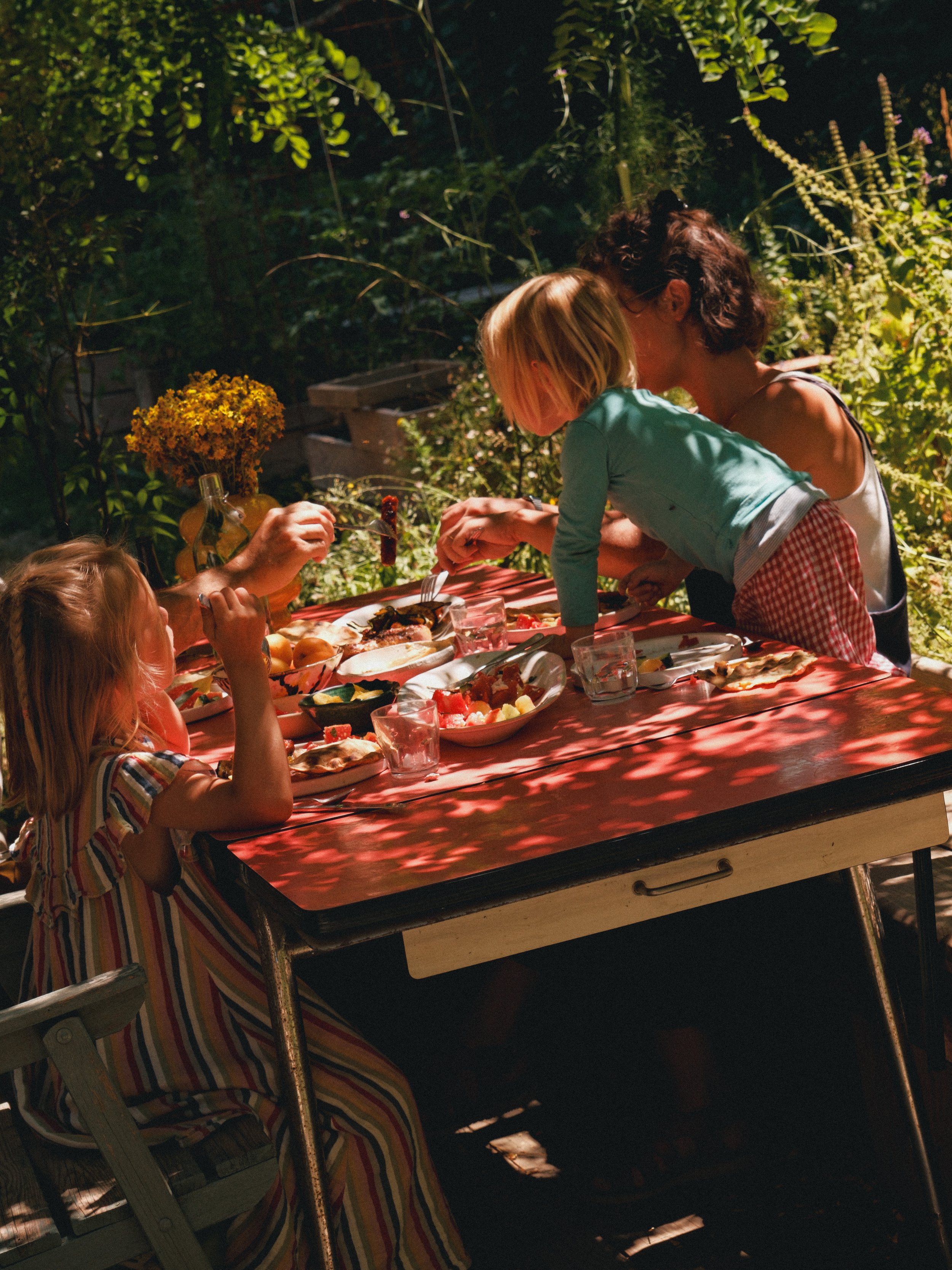Une famille d'adultes et d'enfants rassemblés autour d'une table extérieure, partageant un repas en soirée, sous des arbres avec une lumière tamisée.