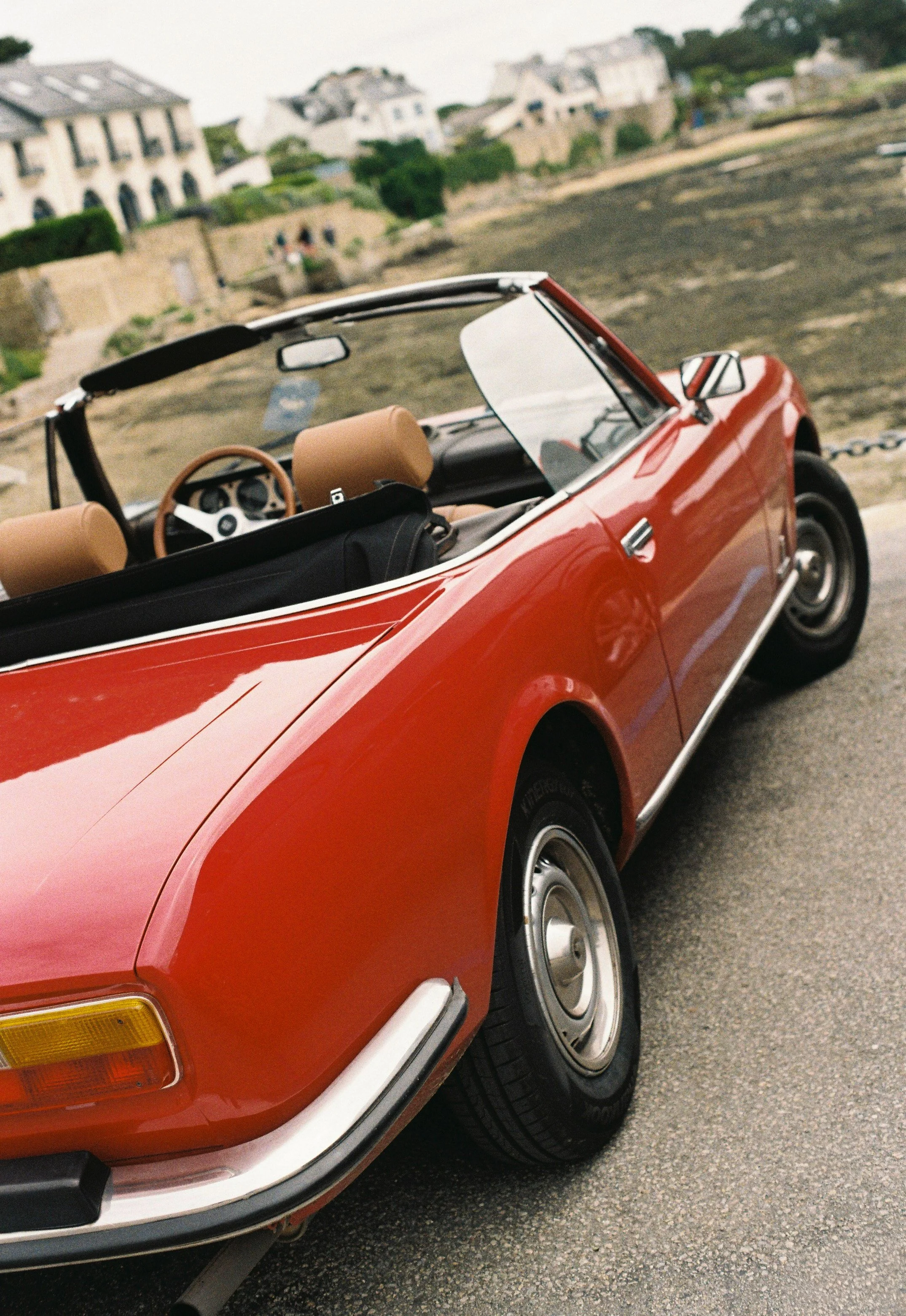 Voiture de couleur rouge avec toit rétractable et intérieur en cuir beige, stationnée sur une route près de maisons en hauteur.