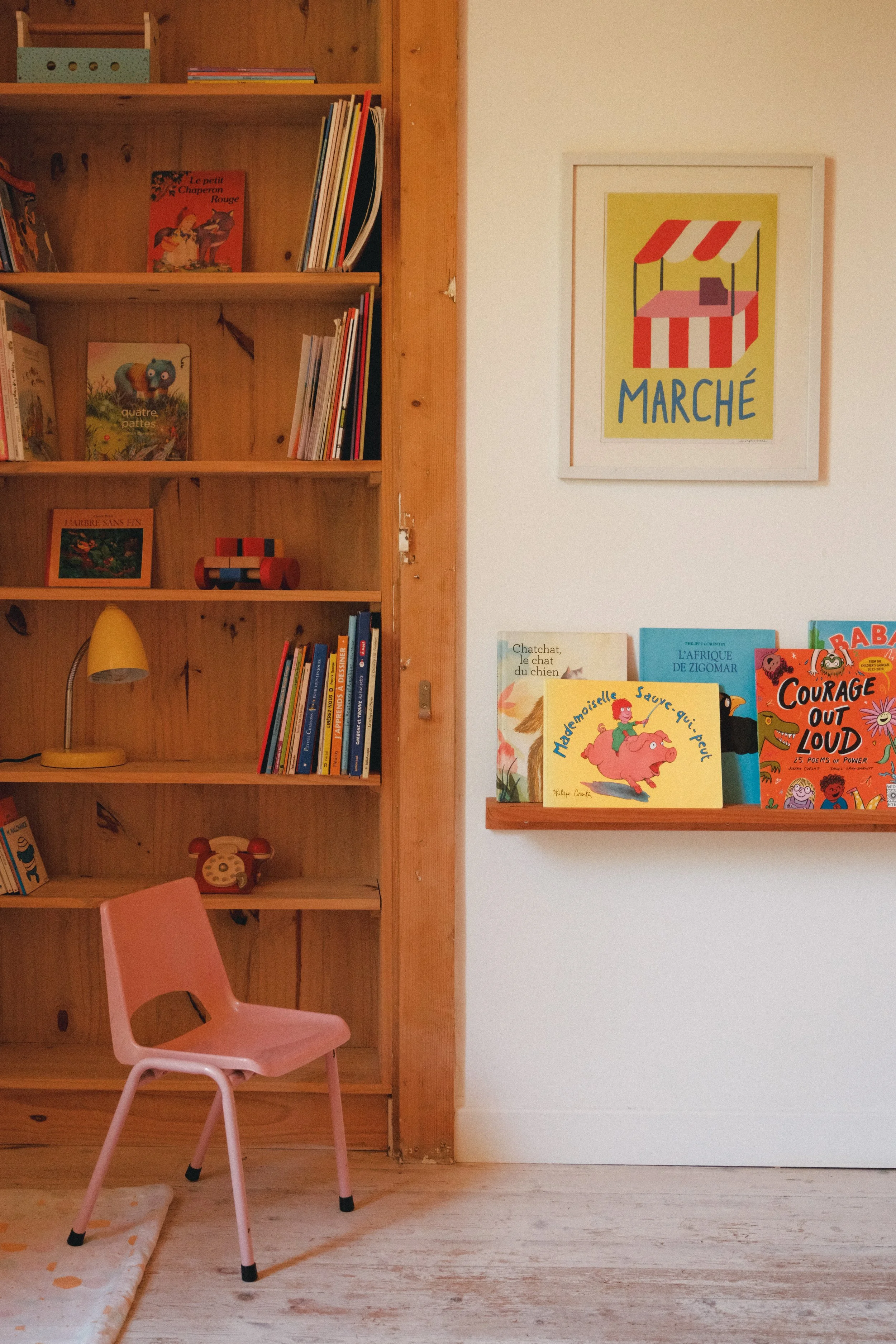 Une étagère en bois avec des livres pour enfants, un cadre avec une affiche de marché, et un fauteuil rose à côté d'un tapis avec un motif de pois. Le sol est en bois clair.