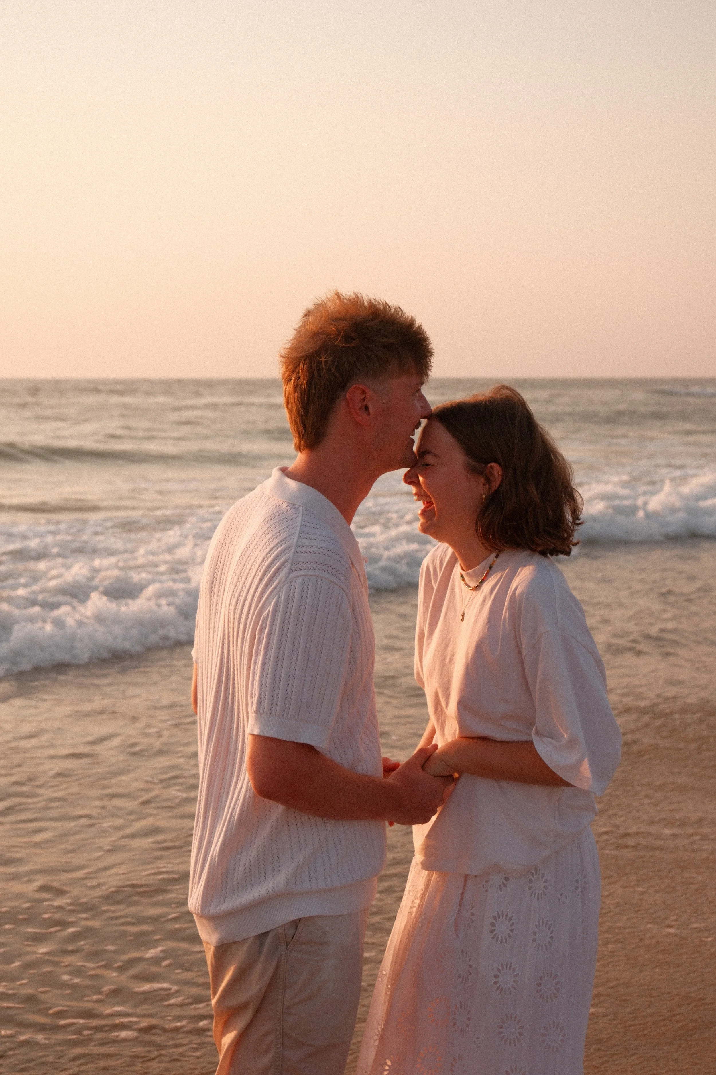 Un couple souriant devant la mer au coucher du soleil, se tenant la main et se regardant tendrement.