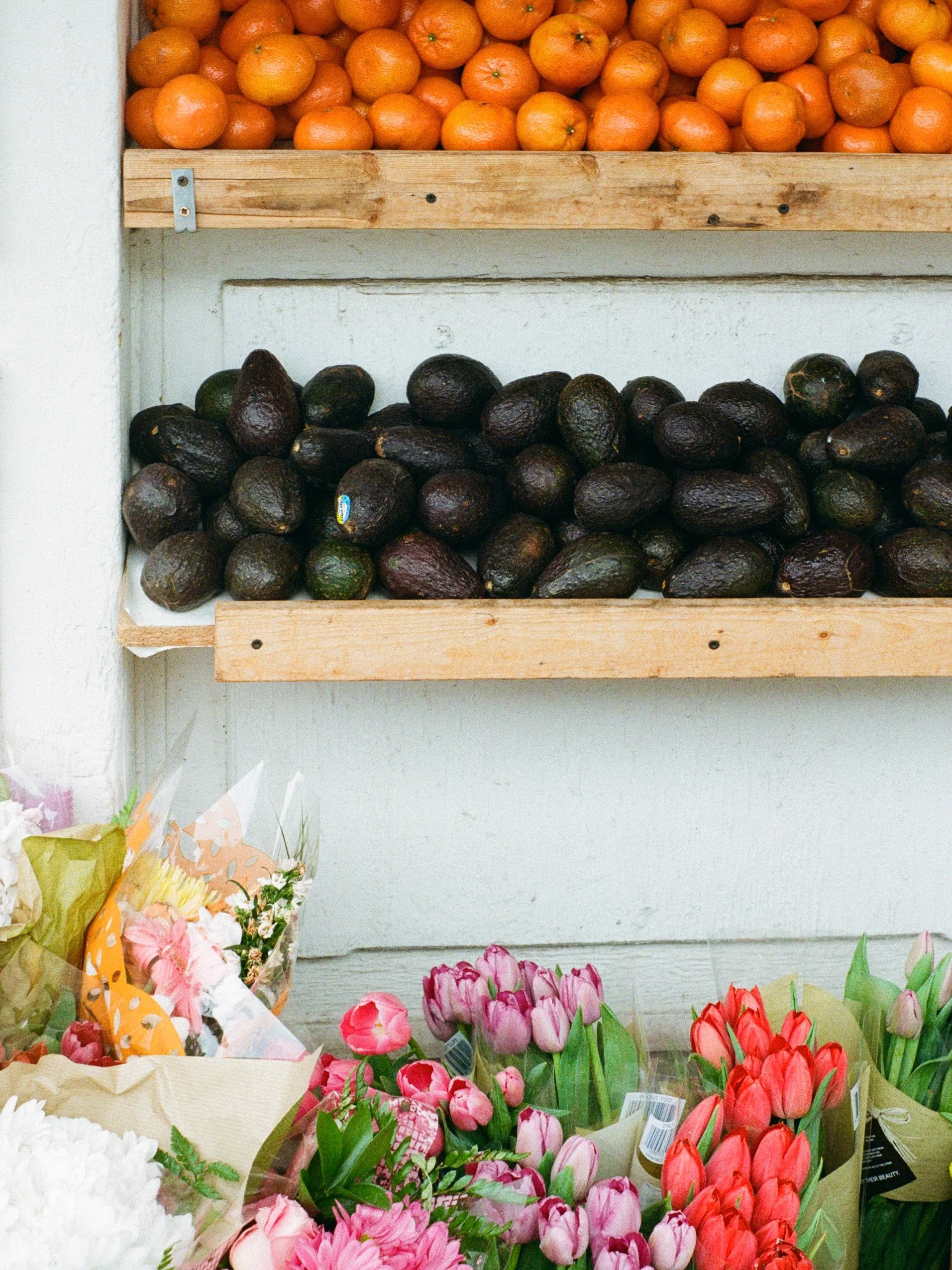 Une étagère avec des mandarines au-dessus et des avocats en dessous, entourés de bouquets de fleurs colorées.