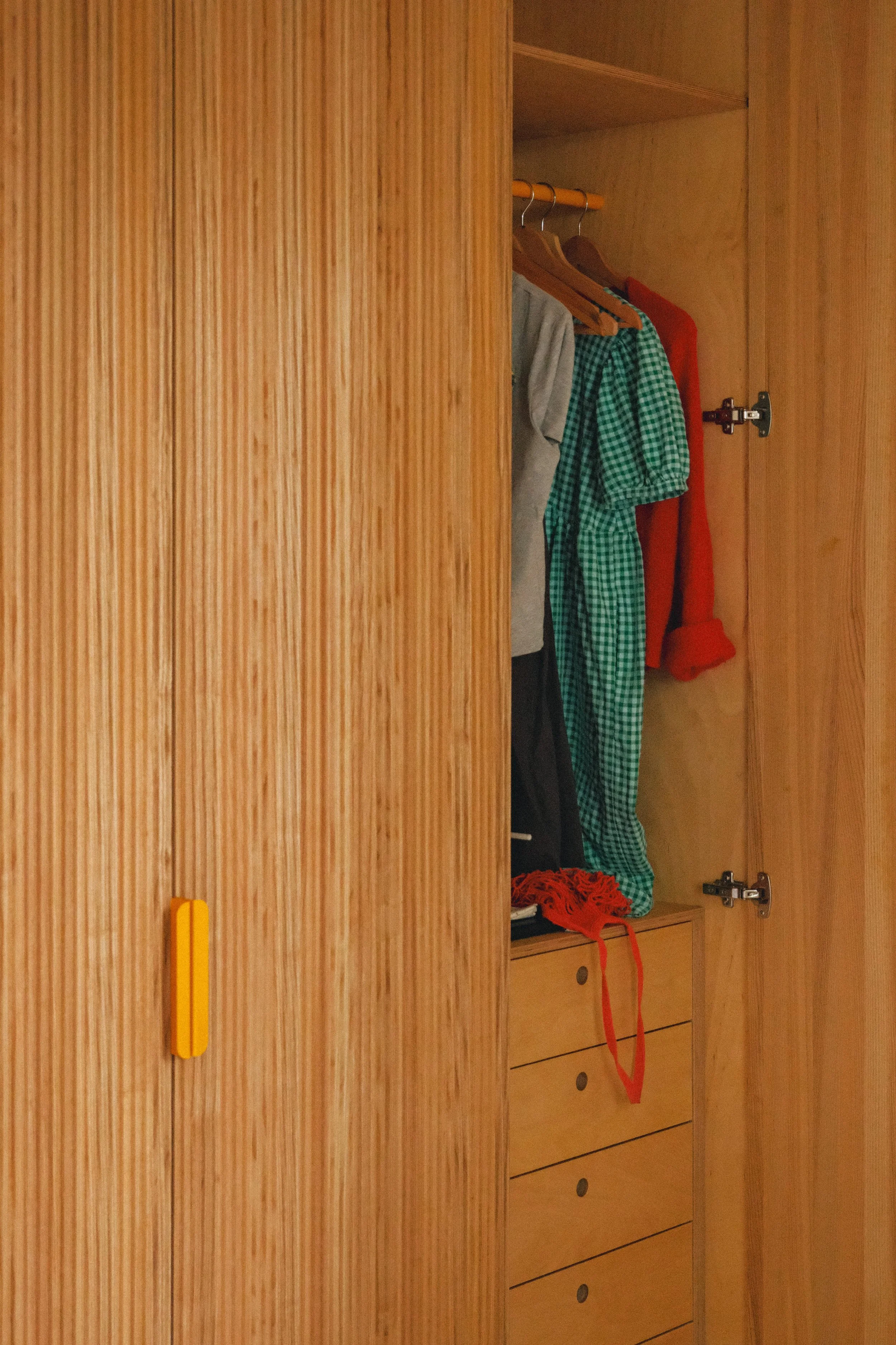 Un placard en bois avec quelques vêtements suspendus à l'intérieur, dont un cardigan gris, une chemise à carreaux vert et blanc, un pull rouge, et une paire de chaussettes ou un vêtement rouge déplié sur le tiroir inférieur.