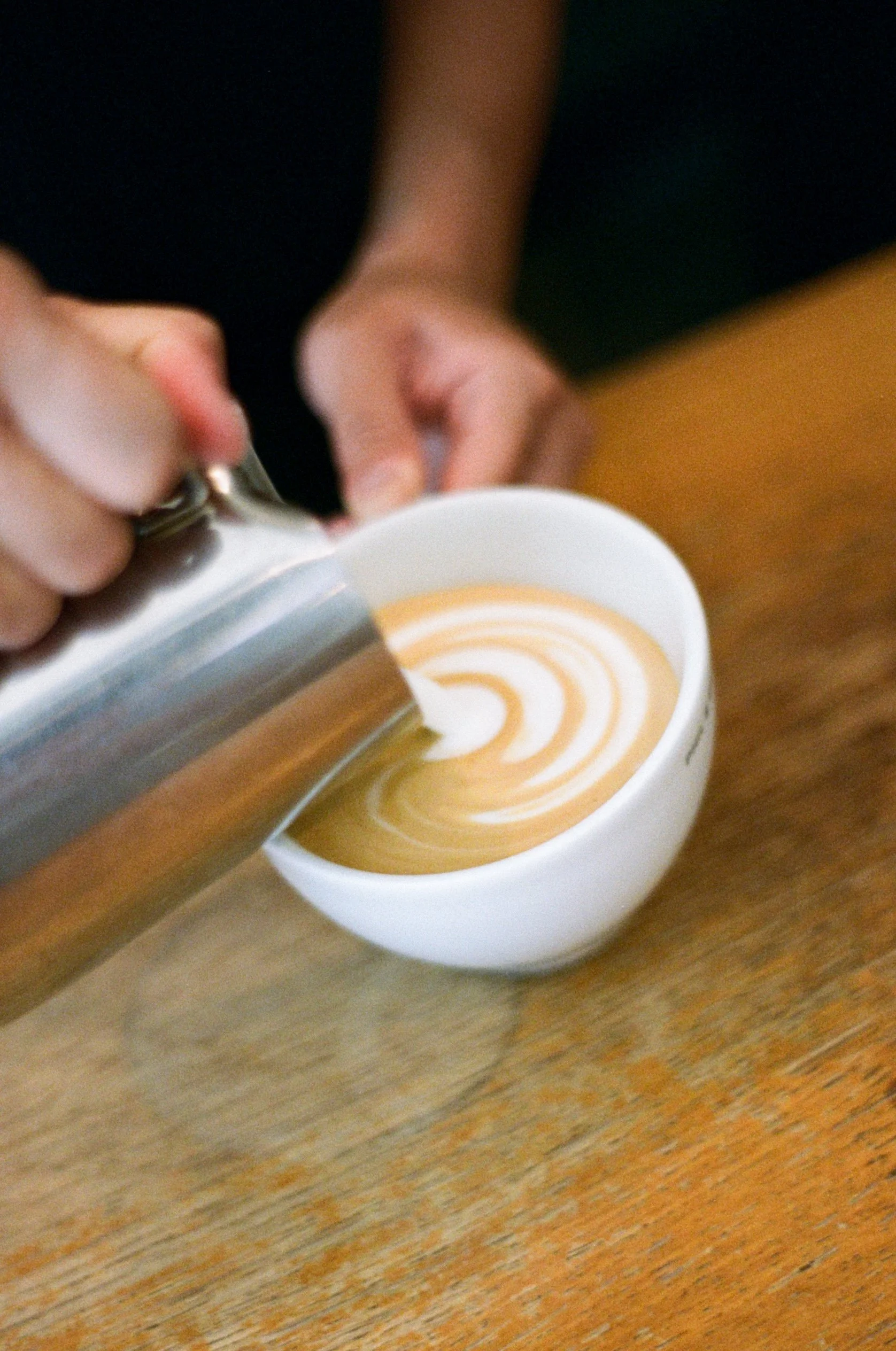Une personne verse un lait chaud ou un autre liquide dans une tasse à latte avec un motif en spirale, posé sur une surface en bois.