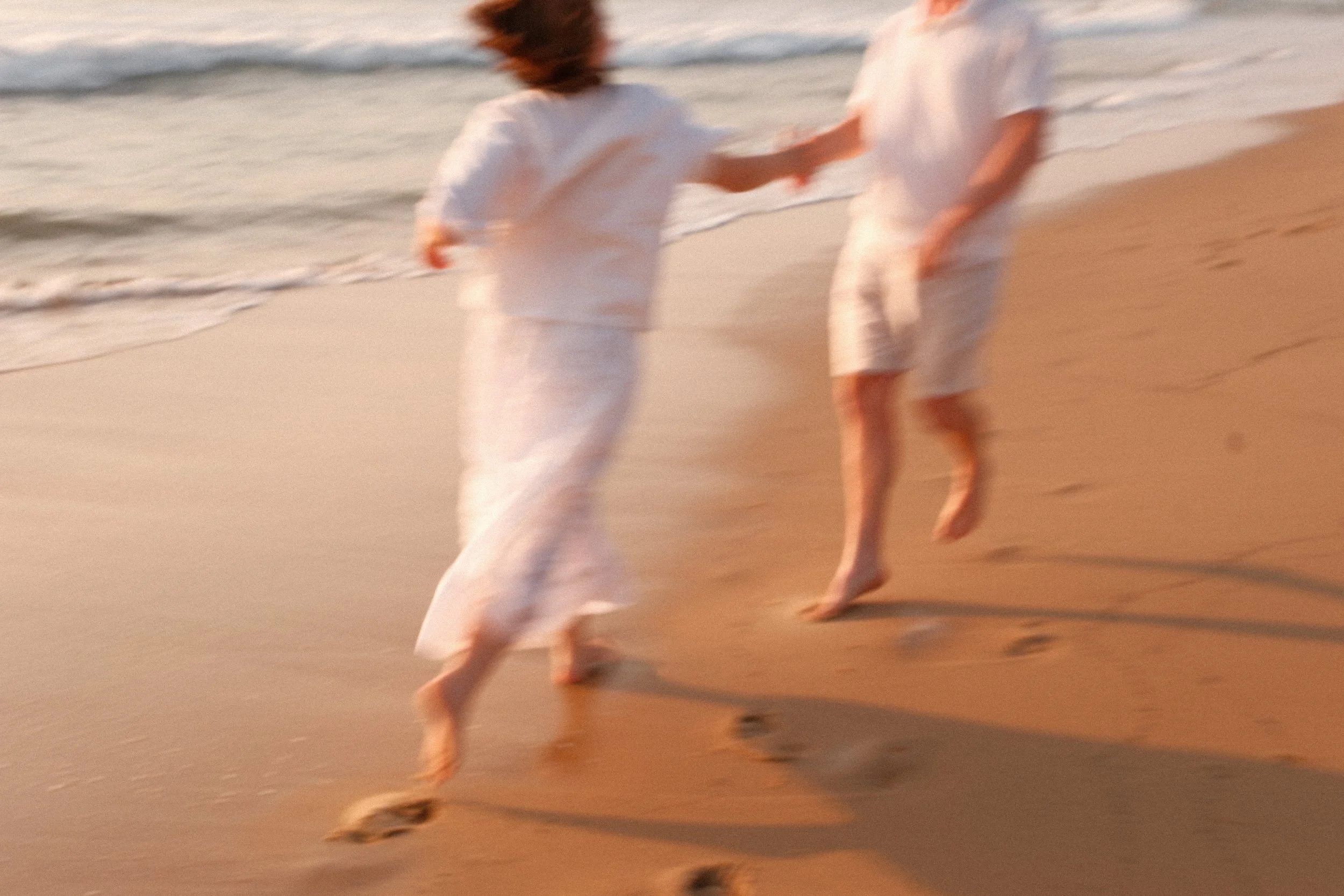 Deux personnes en vêtements blancs marchant ou courant pieds nus sur la plage au coucher du soleil.