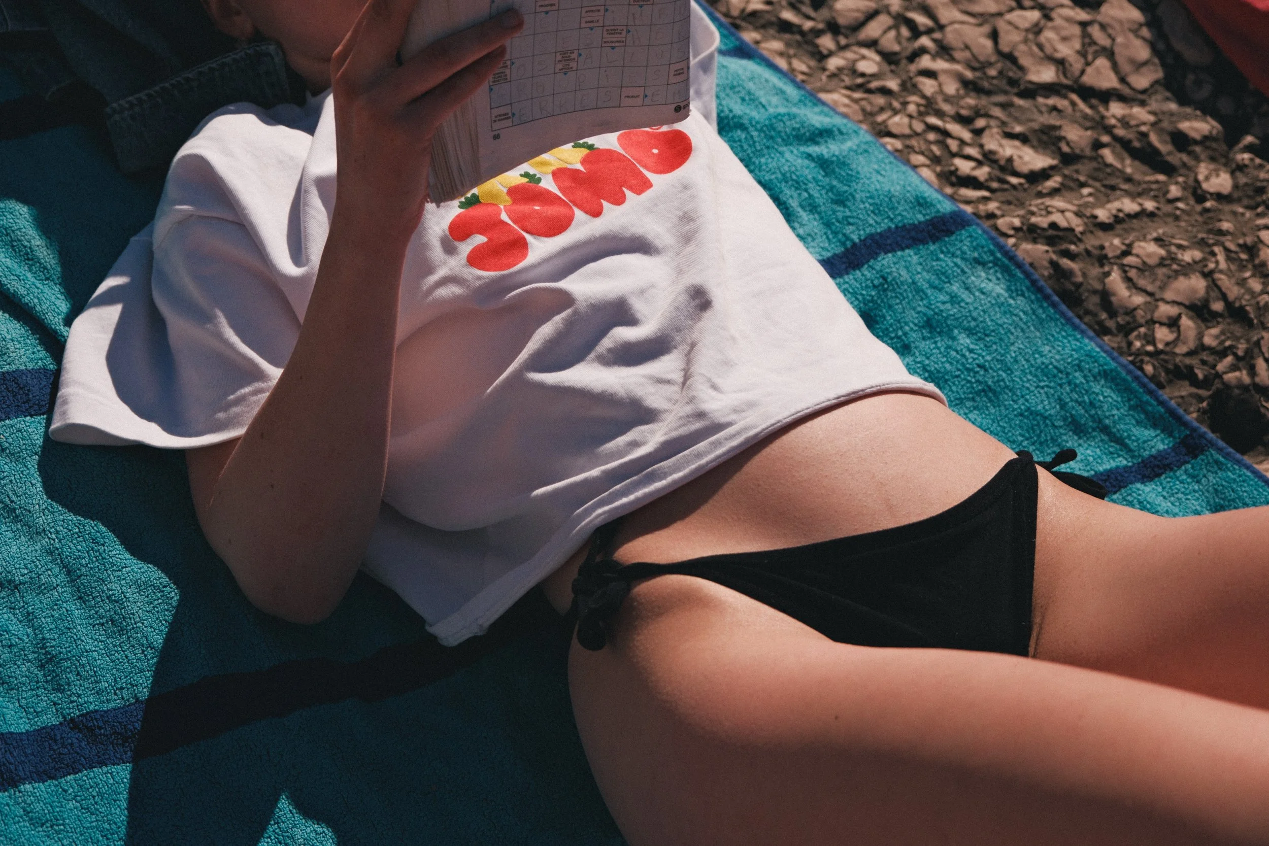 Une personne allongée sur une serviette turquoise, portant un t-shirt blanc avec le mot 'Famos' écrit dessus et des dessins de fraises, tenant un billet ou un document.