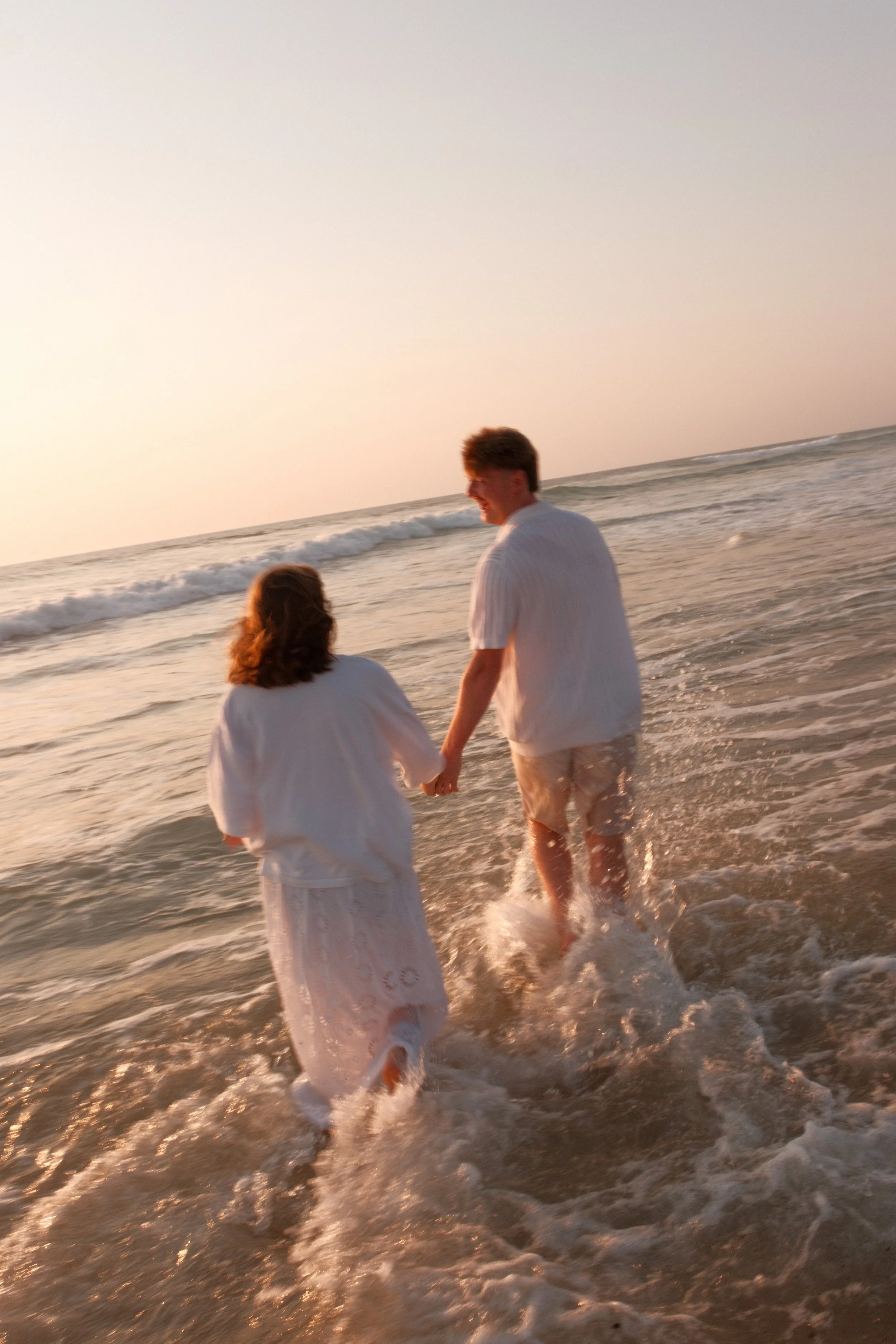 Deux personnes marchent main dans la main dans la mer au coucher du soleil, portant des vêtements blancs, ambiance chaleureuse et romantique.